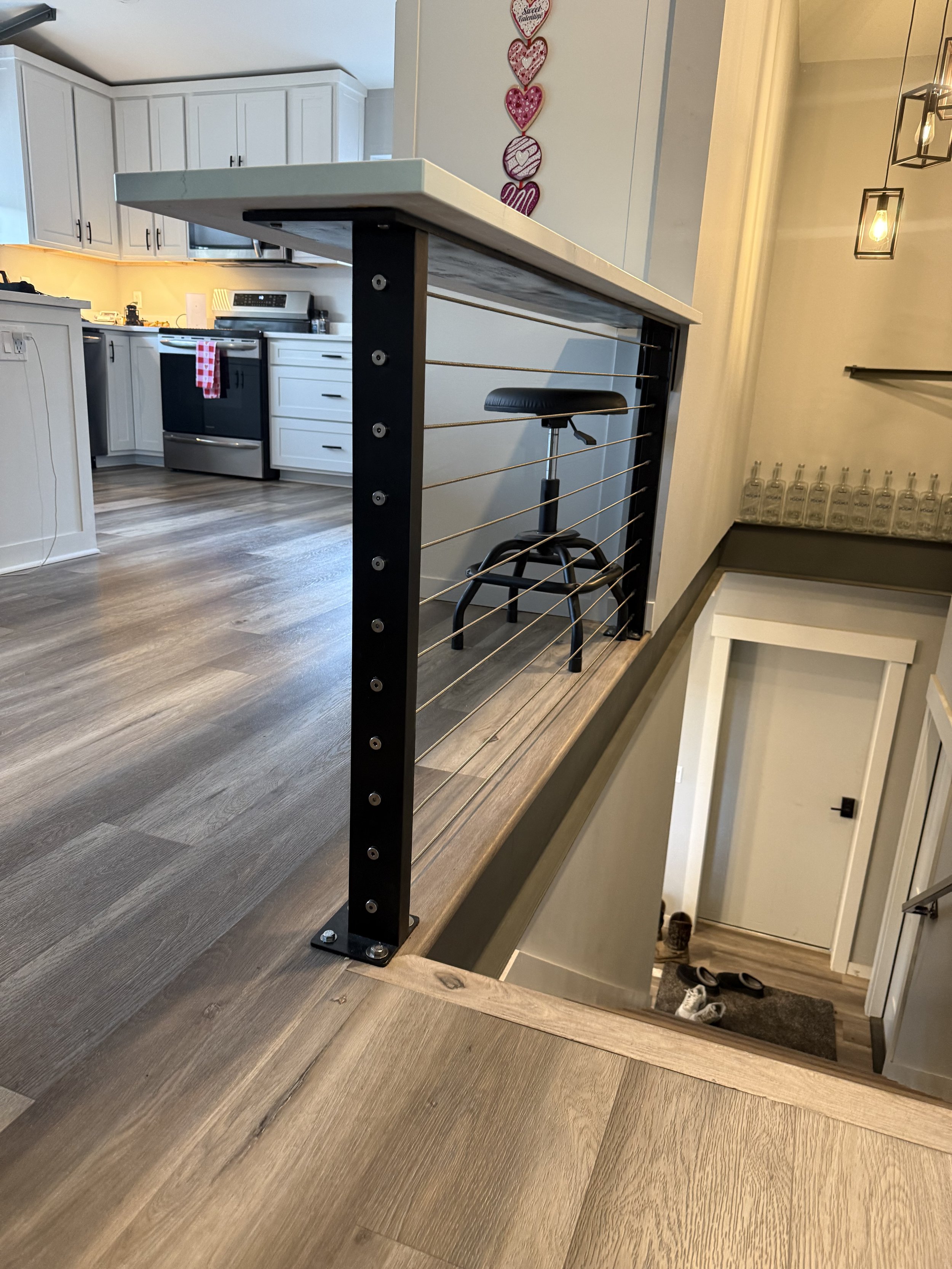 View of a kitchen with a wooden floor, white cabinets, and a black stove. A bar with a metal railing and a black stool overlooks a staircase going down.