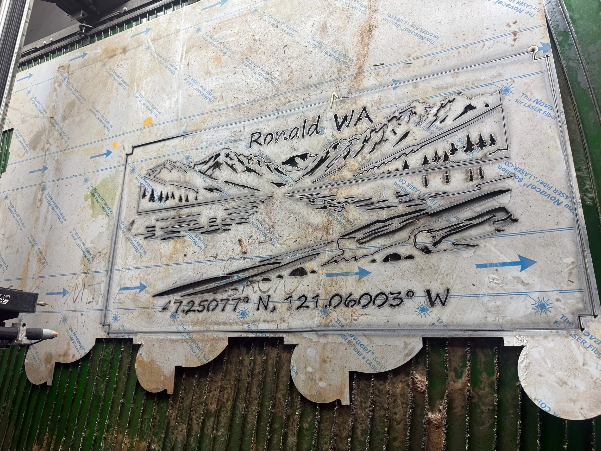 Metal cutout sign with mountain landscape and houses, inscribed with 'Ronald WA' at the top, and GPS coordinates at the bottom.