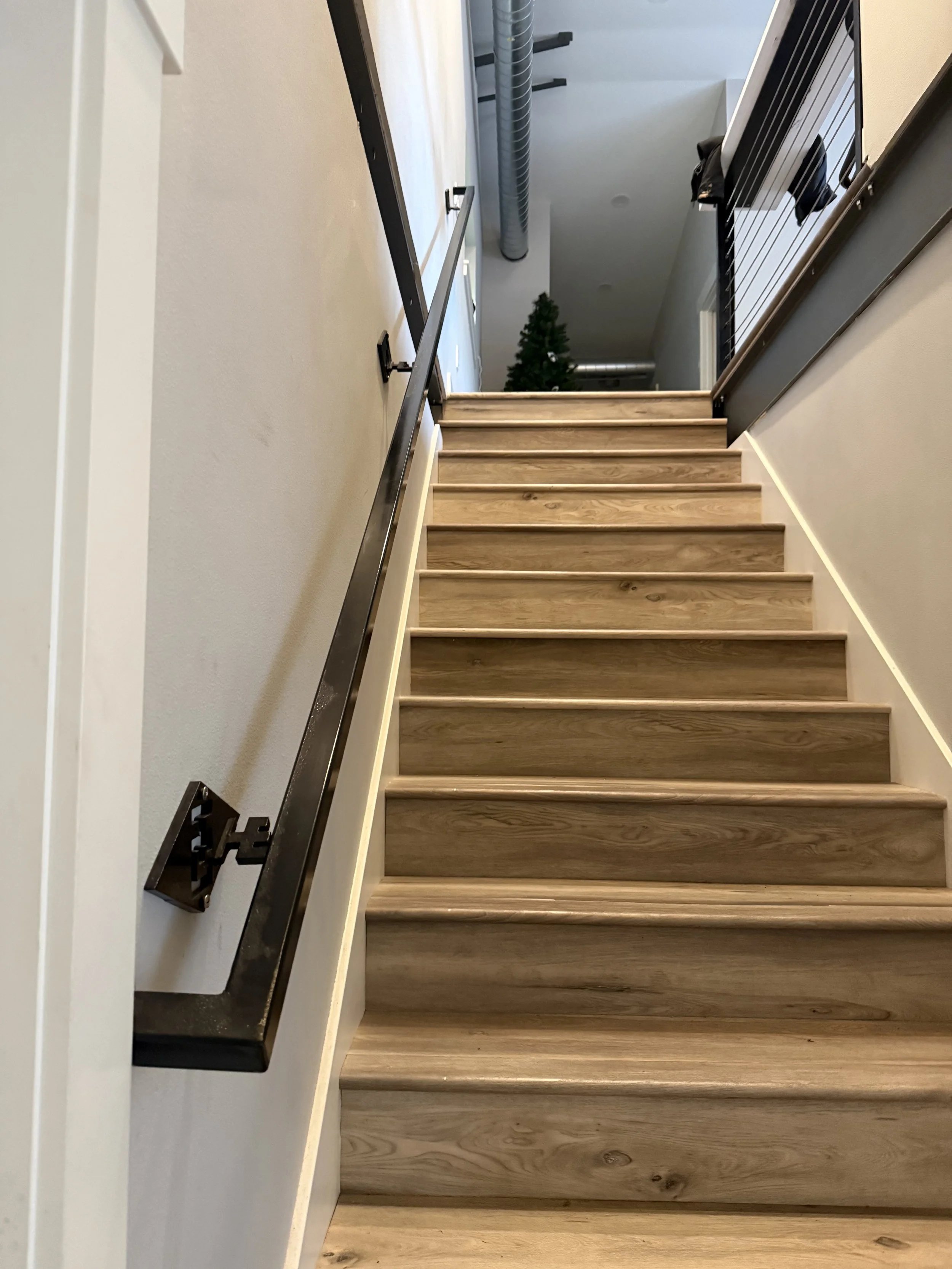 Wooden staircase viewed from below, with white walls and metal handrails on both sides, leading up to a hallway with a Christmas tree in the background.