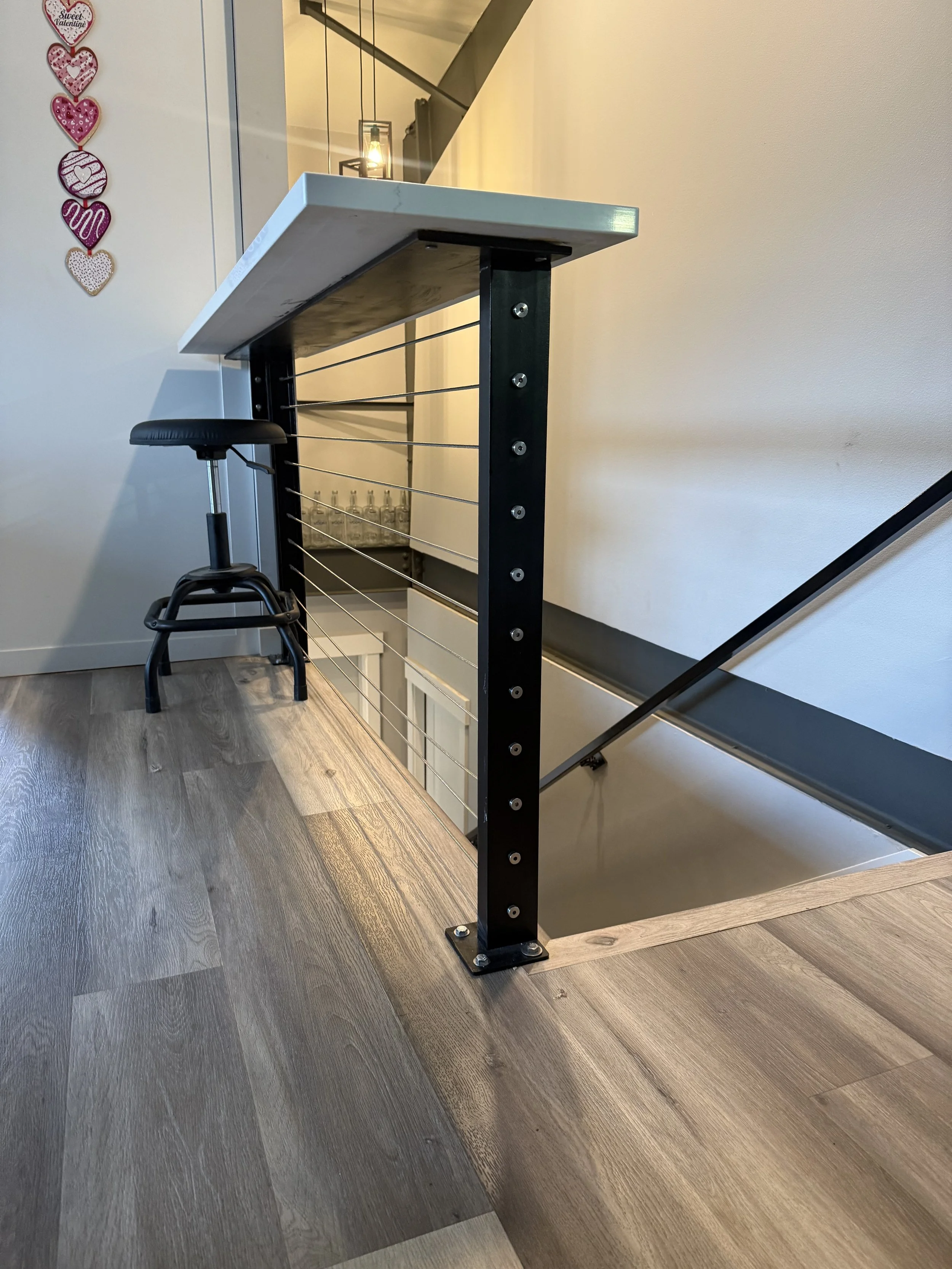 A modern staircase railing with black metal posts and horizontal metal wires, topped by a white marble or stone handrail, situated next to a staircase with wood flooring.