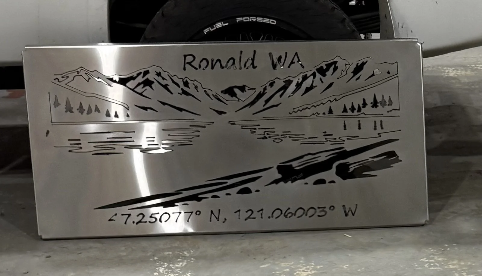 Metal plaque with a mountain, lake, trees, and road design, inscribed with "Ronald WA" and GPS coordinates "47.25077° N, 121.06003° W".