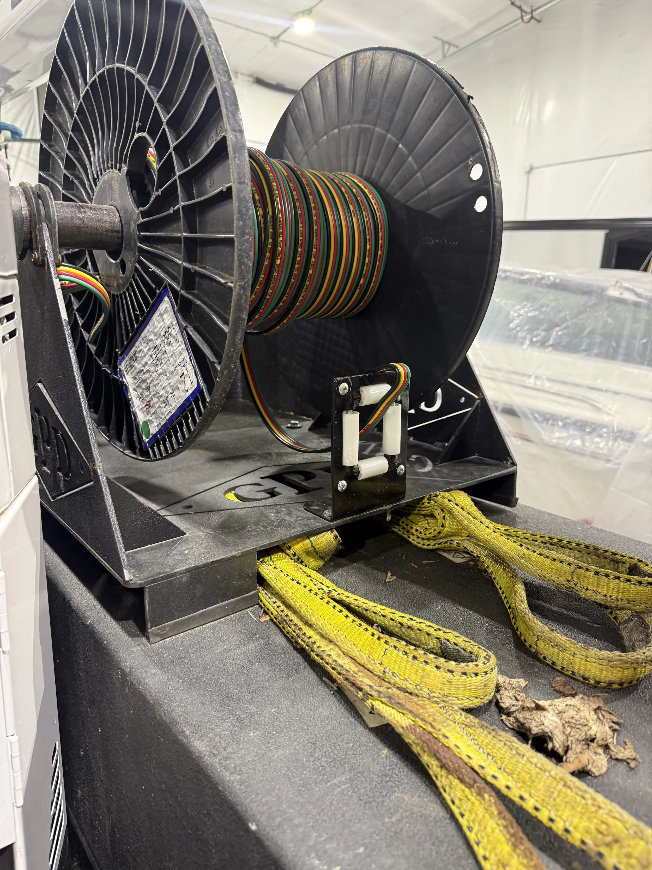 A large industrial reel of multicolored electrical wire mounted on a black stand, with yellow tow straps on a black surface and a white fabric background.
