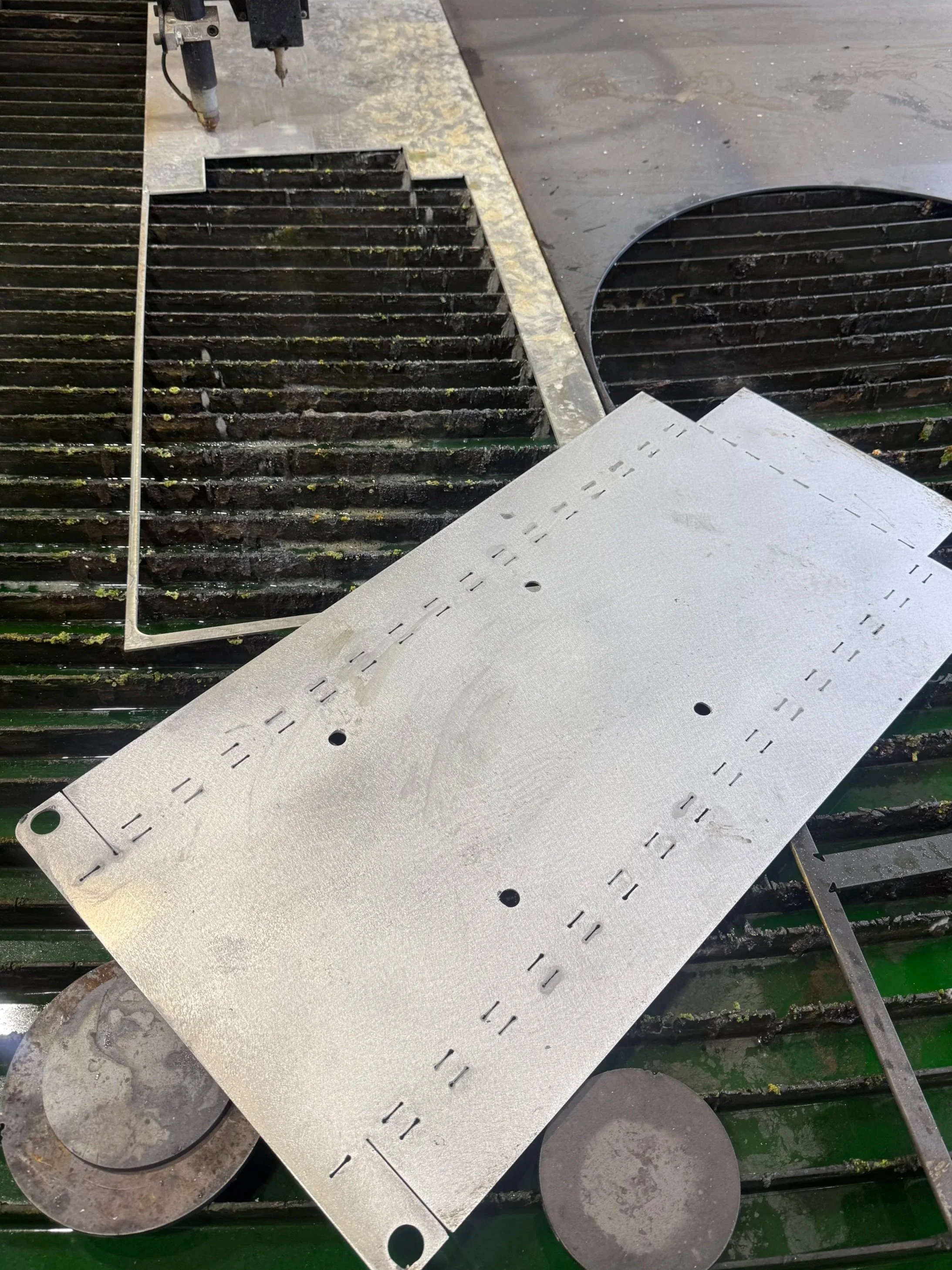 Metal sheet with evenly spaced slots and circular holes, placed on top of a grill at a workshop or industrial setting.