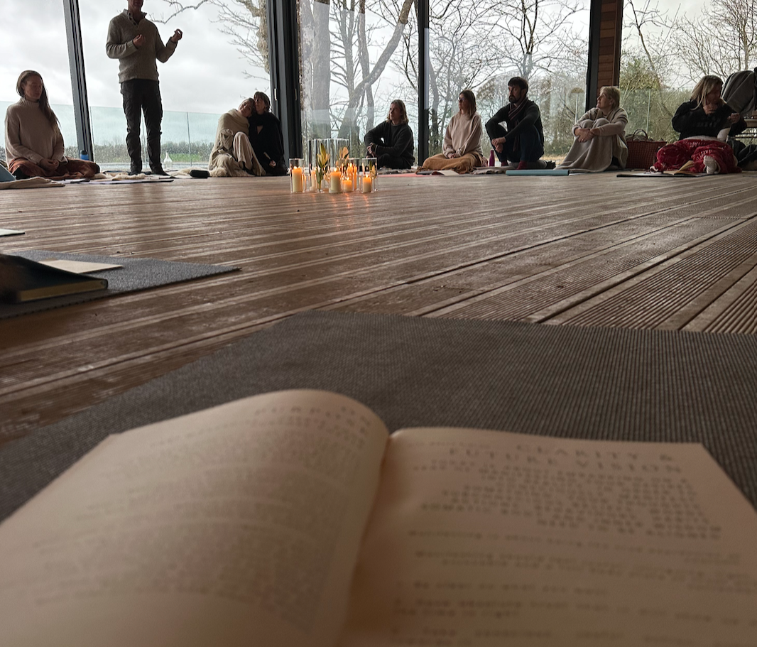 A group of people sitting on the floor in a semi-circle inside a room with large windows, candles in the center, and scenic outdoor views. Someone is standing and speaking to the group, with a book open in the foreground.