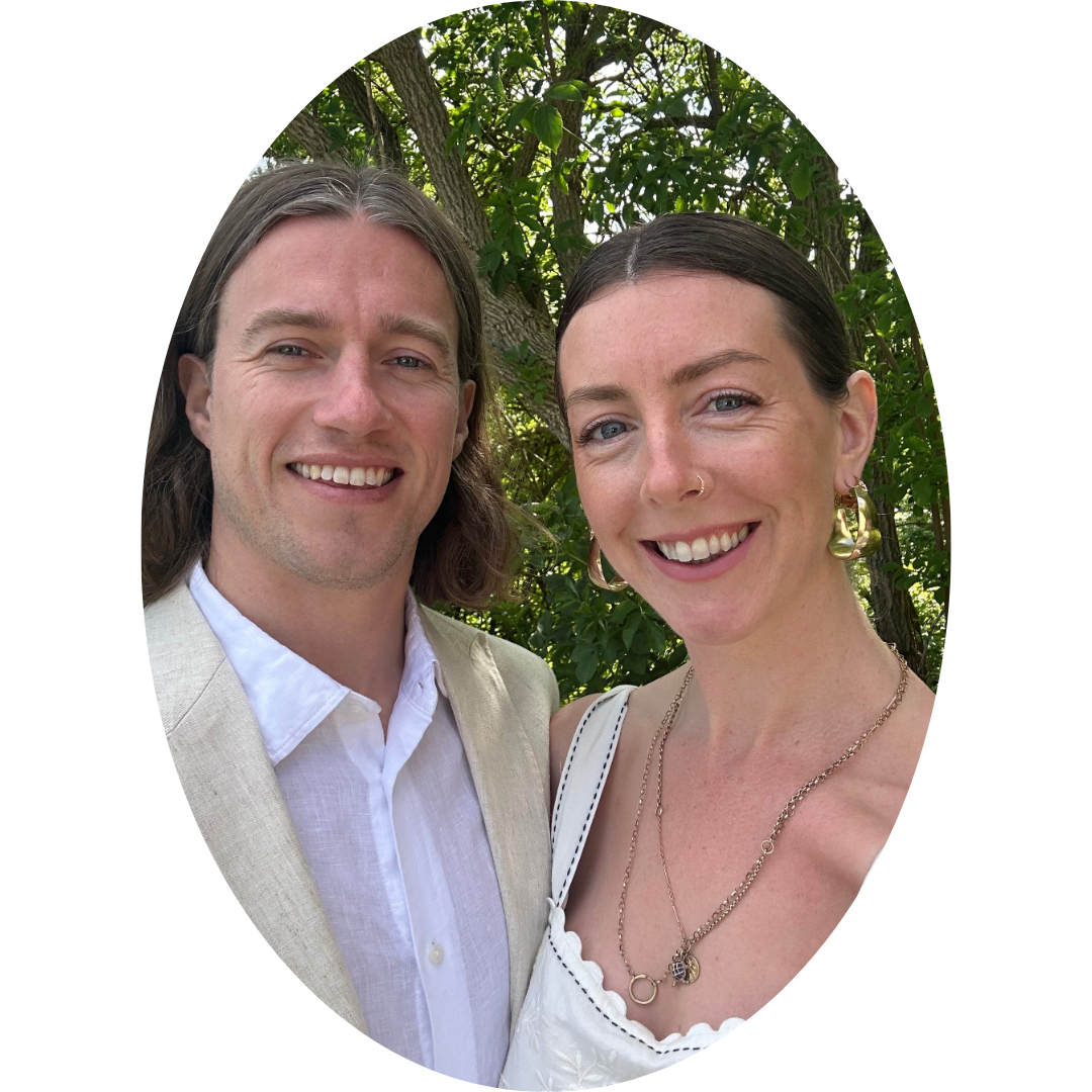 A man and a woman smiling outdoors in front of green trees.