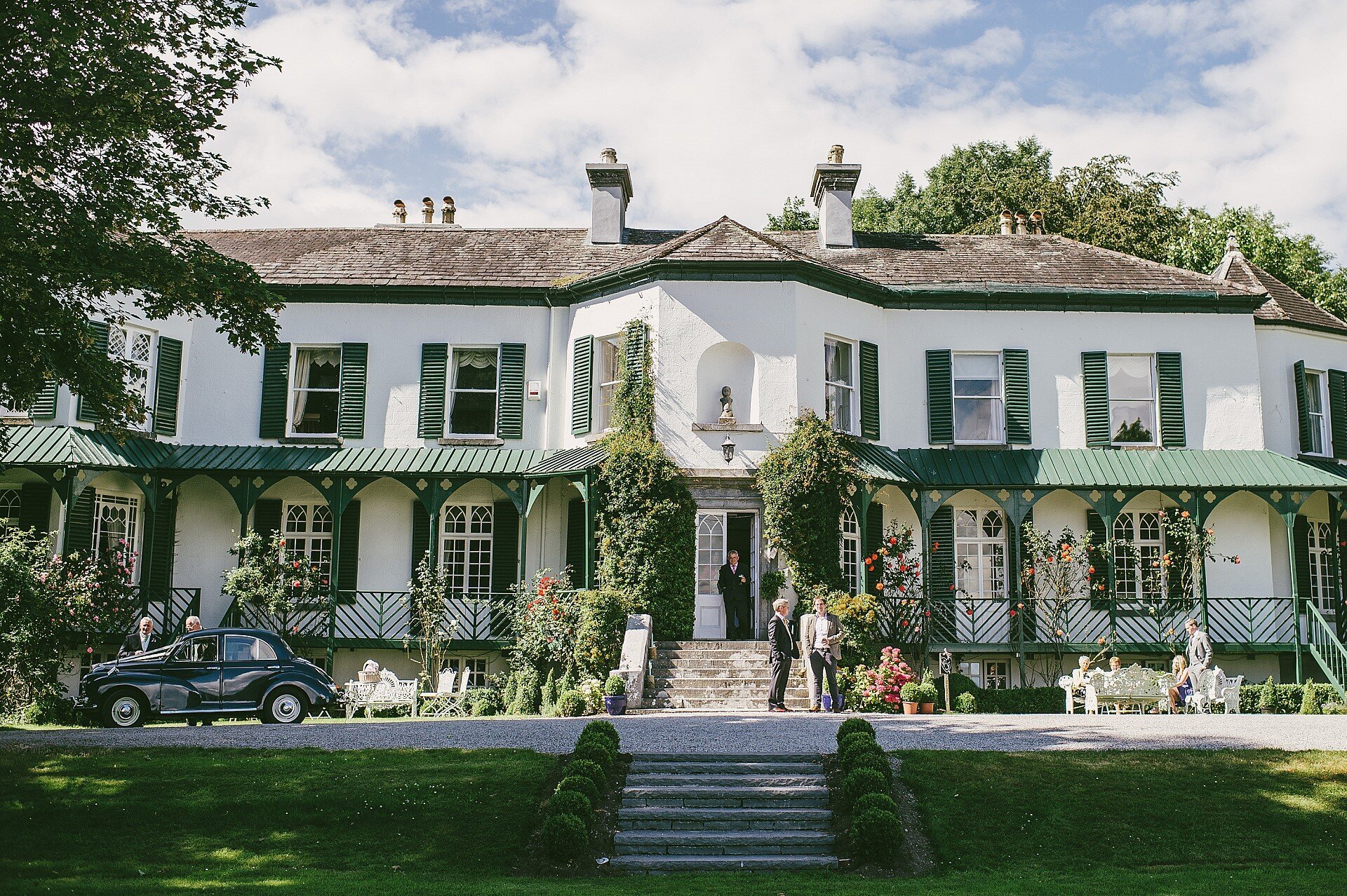 Large white mansion with green shutters and awnings, surrounded by garden with people, vintage black car, benches, and potted plants.