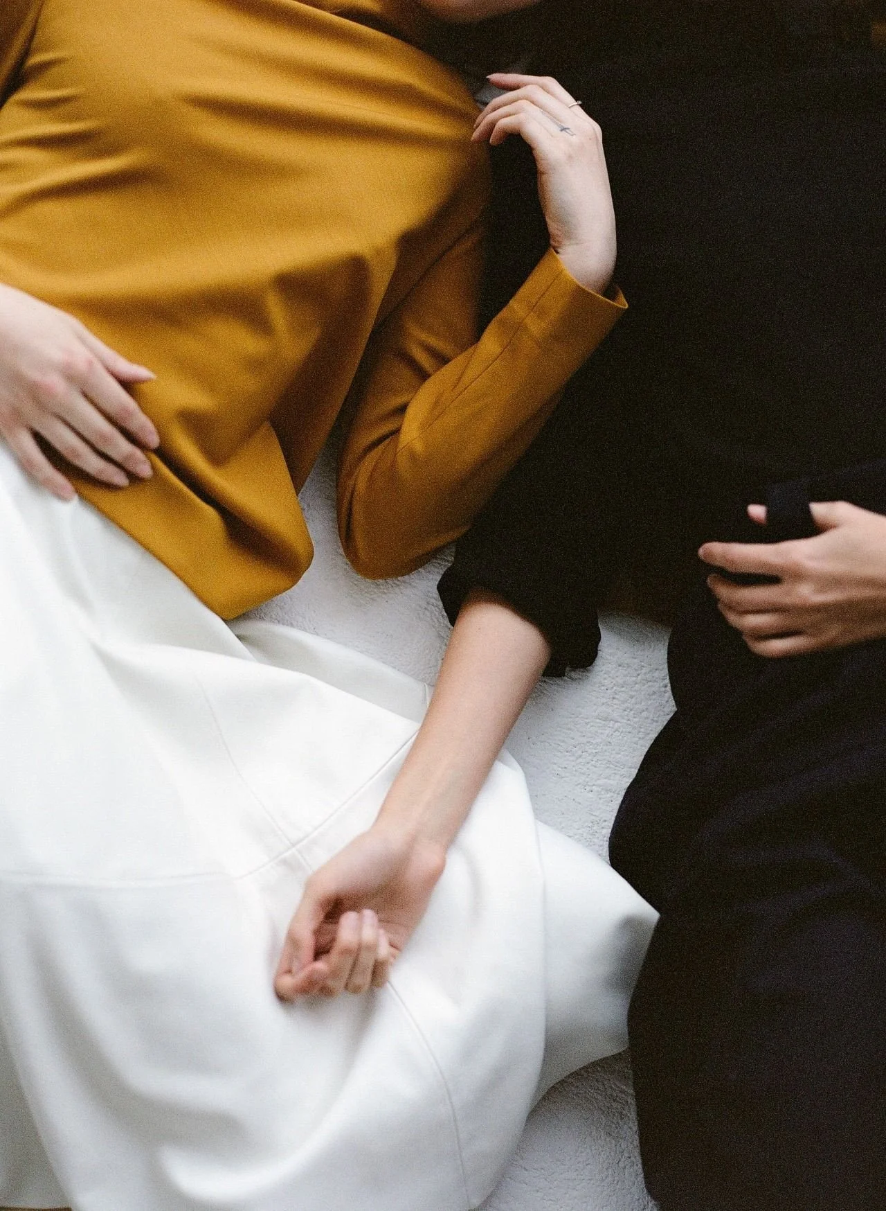 Close-up of two people lying on a white surface, one wearing a mustard yellow jacket and the other in black, holding hands.