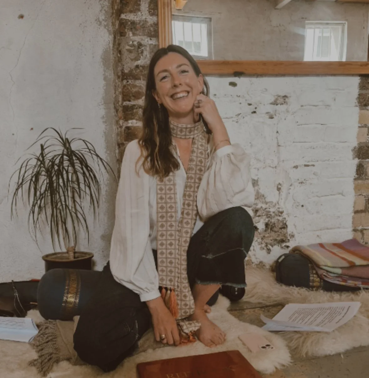 A woman with long brown hair kneeling on a furry rug, smiling and touching her face. She wears a patterned scarf, white blouse, and black ripped jeans. The background shows a rustic brick and plaster wall, a potted plant, and some papers and bags on the floor.