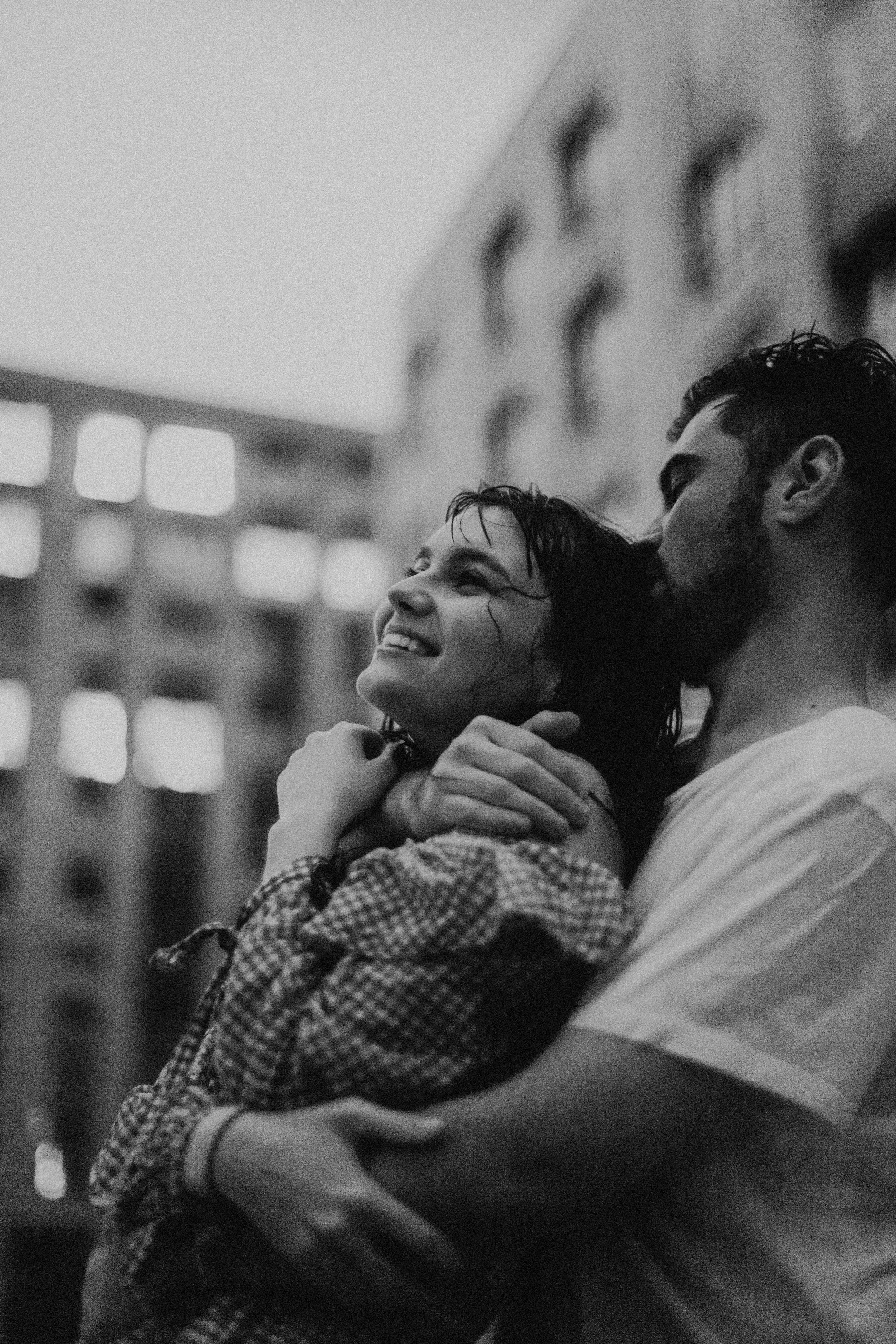 A black and white photo of a man holding a woman, both smiling, with buildings in the background.