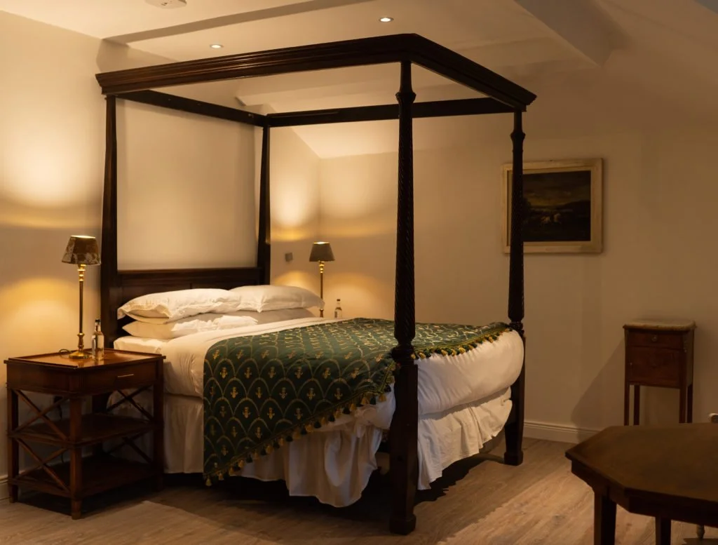 A bedroom with a dark wooden four-poster bed, white linens, and a decorative green and gold bedspread. There are two matching bedside lamps on wooden nightstands and a framed landscape painting on the wall. The room has beige walls and soft lighting.