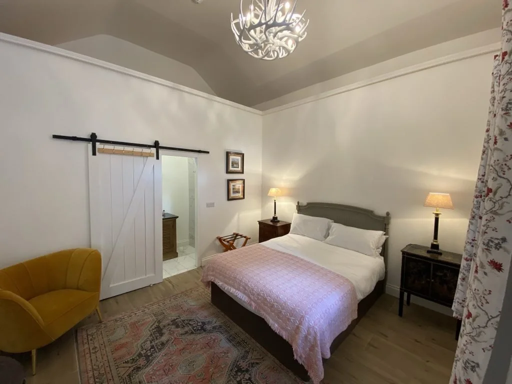 A cozy bedroom with a bed, two nightstands with lamps, a yellow armchair, a patterned rug, a decorative light fixture on the ceiling, and a sliding barn door leading to a bathroom.