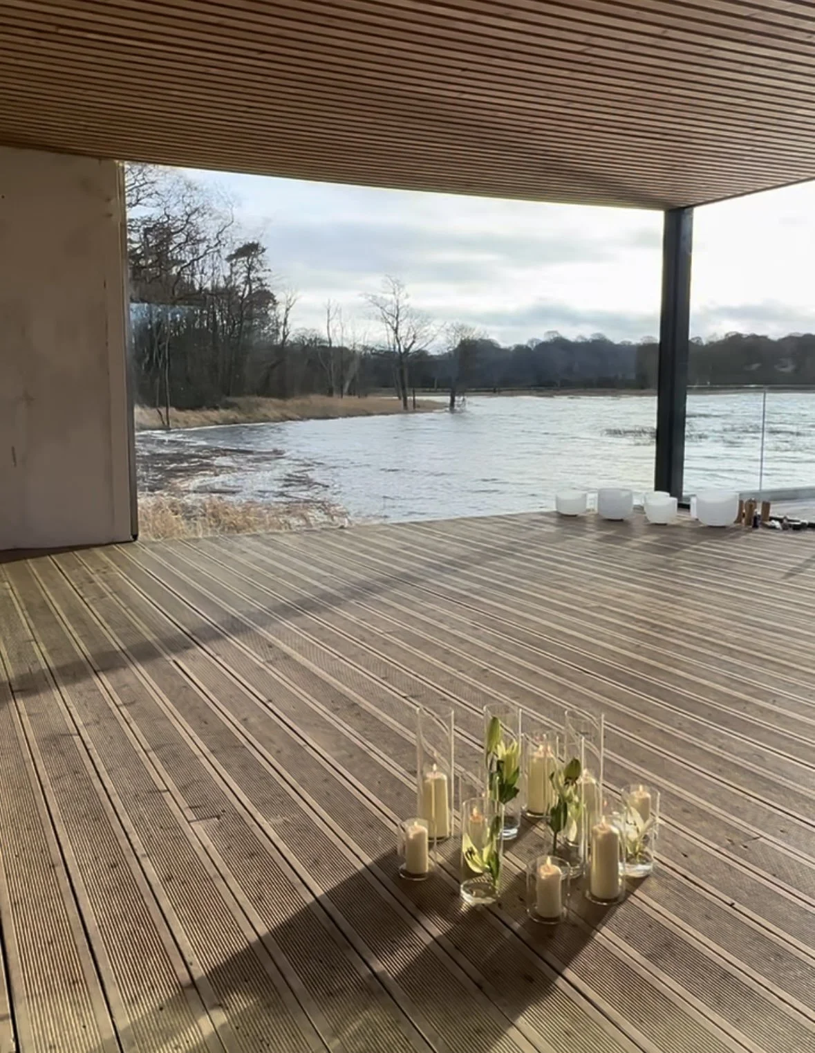An interior view of a modern lakeside room with large open windows, wooden flooring, and a decorative arrangement of candles and flowers on the floor.