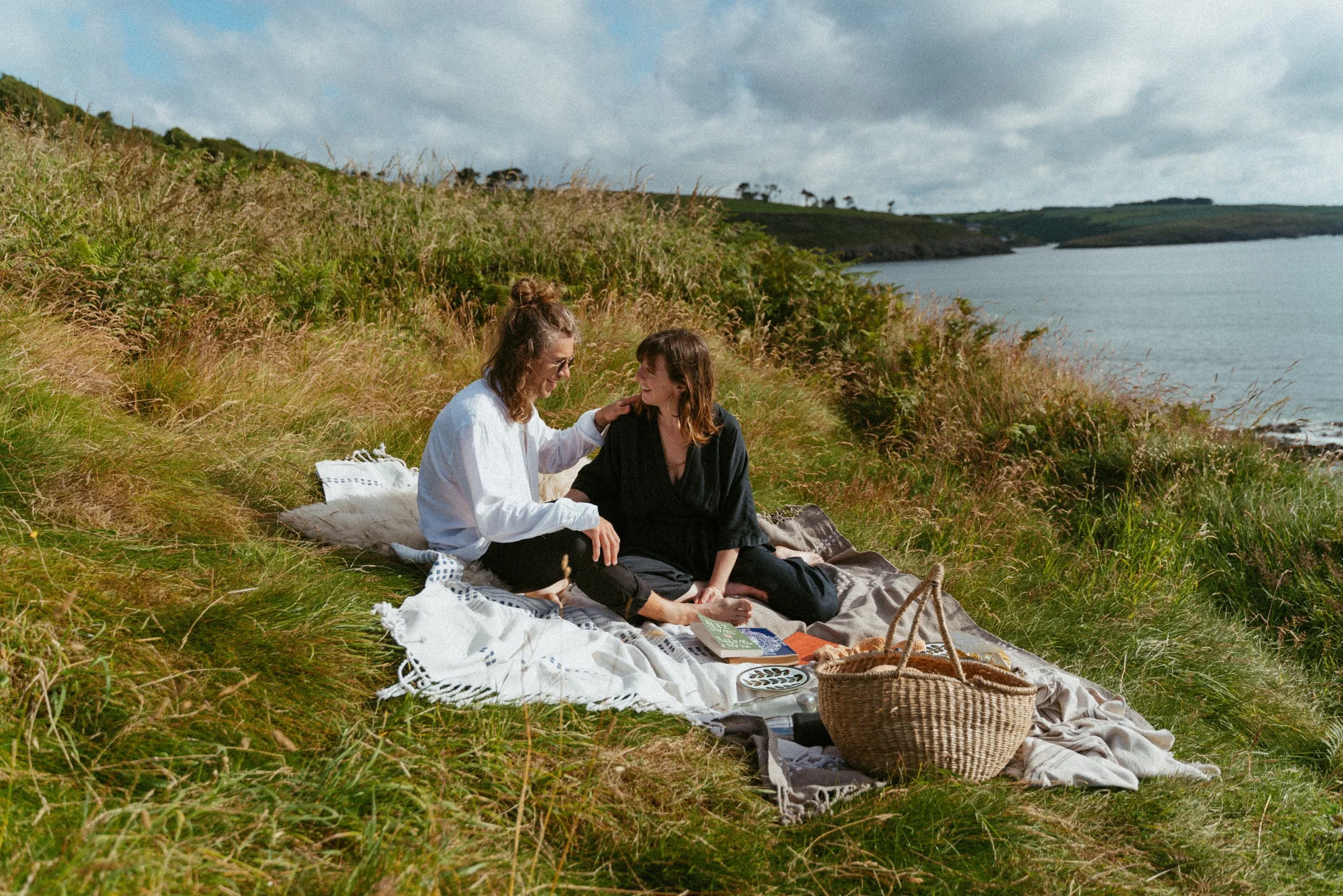 Two women enjoying a picnic on a grassy hillside near a body of water, with a picnic basket and blankets.