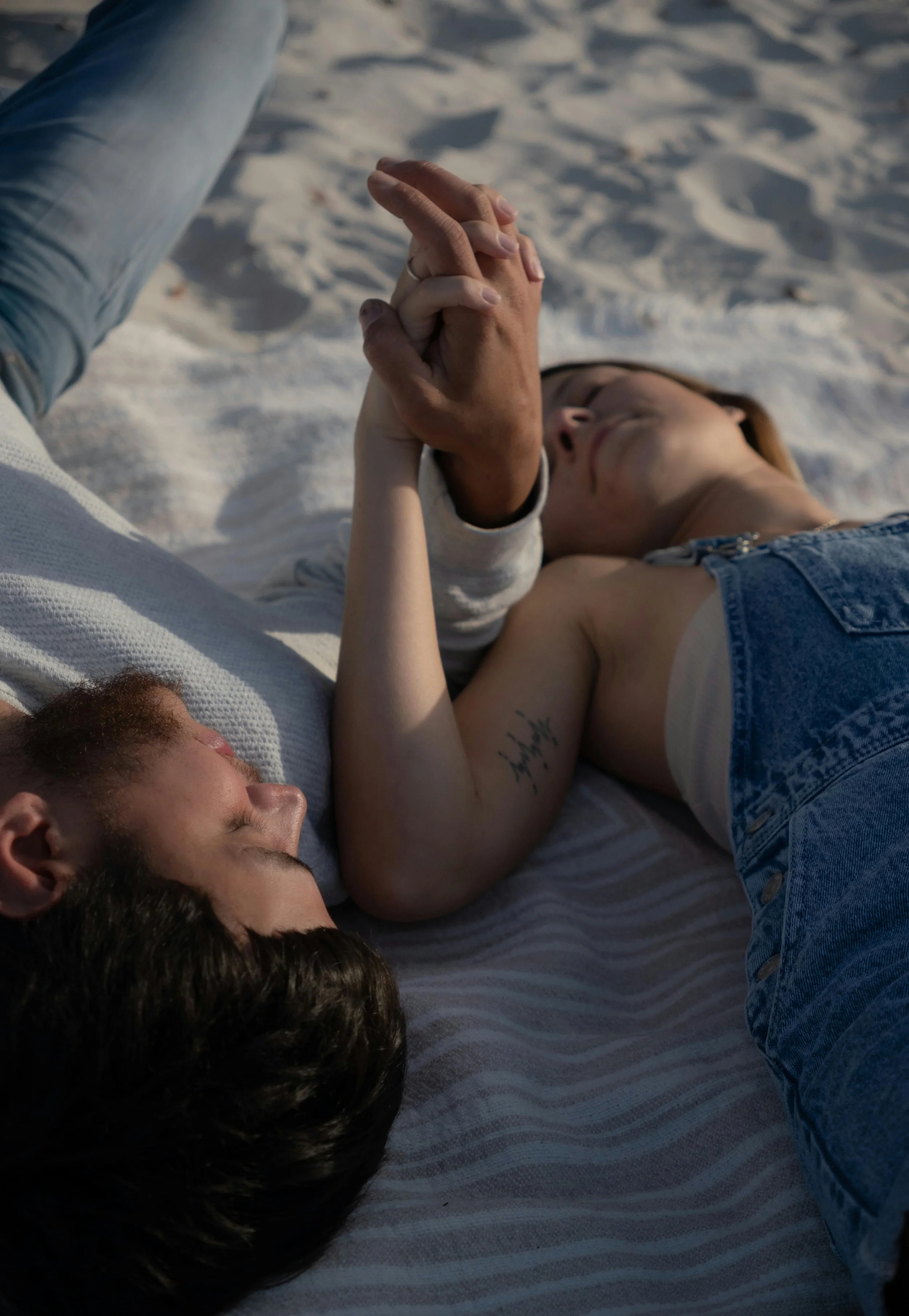 A couple lying on a blanket in a sandy outdoor area, holding hands, with their eyes closed and faces relaxed.