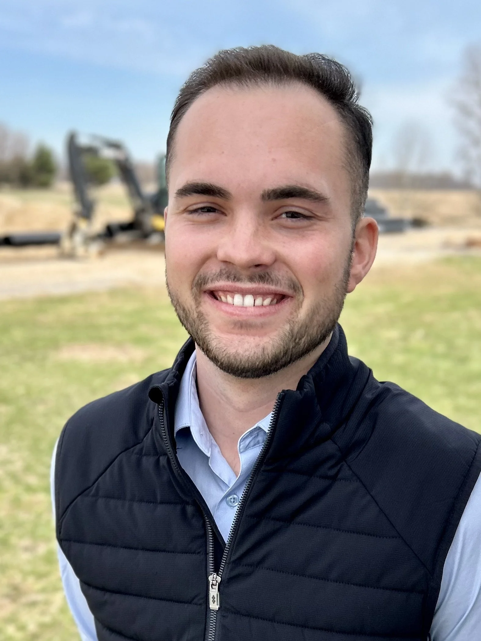A smiling man with short dark hair, a beard, wearing a light blue button-up shirt and a black padded vest standing outdoors on a grassy area, with blurred machinery and trees in the background.
