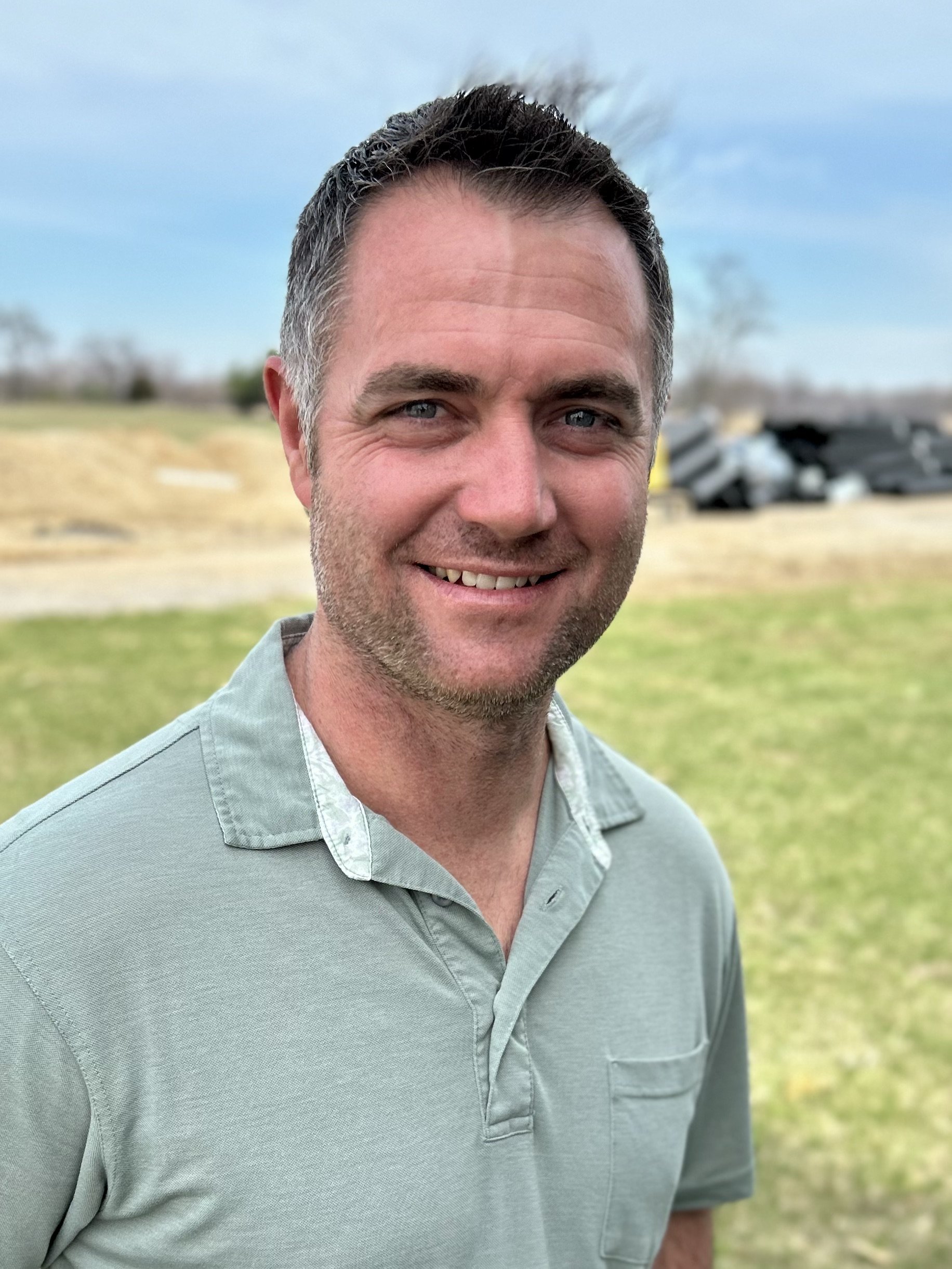 A smiling man with short dark hair, light skin, and wearing a light green collared shirt stands outdoors on grass with a blurred background of trees and sky.