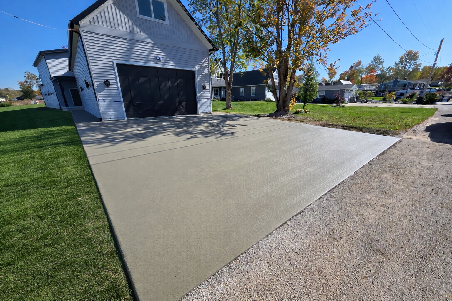 A newly poured concrete driveway extending from a garage to the street, with green grass and trees on either side, in a suburban neighborhood on a sunny day.
