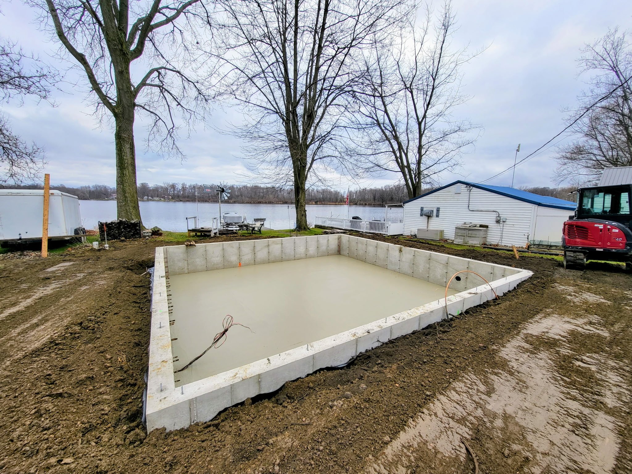 Swimming pool foundation under construction near a lake, with trees and a small building in the background.