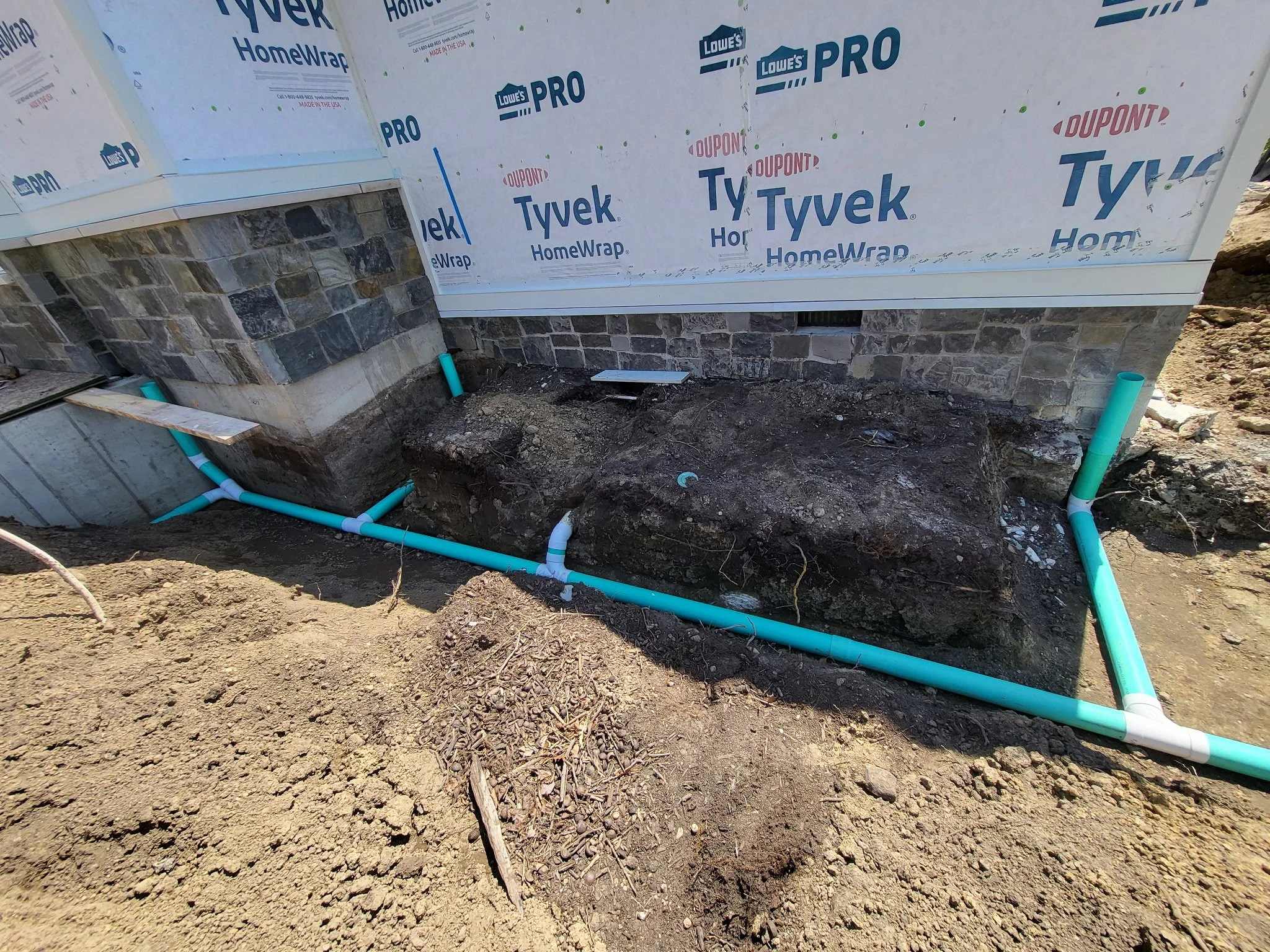 Installation of new plumbing pipes outside a house under construction, with blue and white pipes laid along the foundation near the building's stone wall and wrapped with Tyvek HomeWrap.