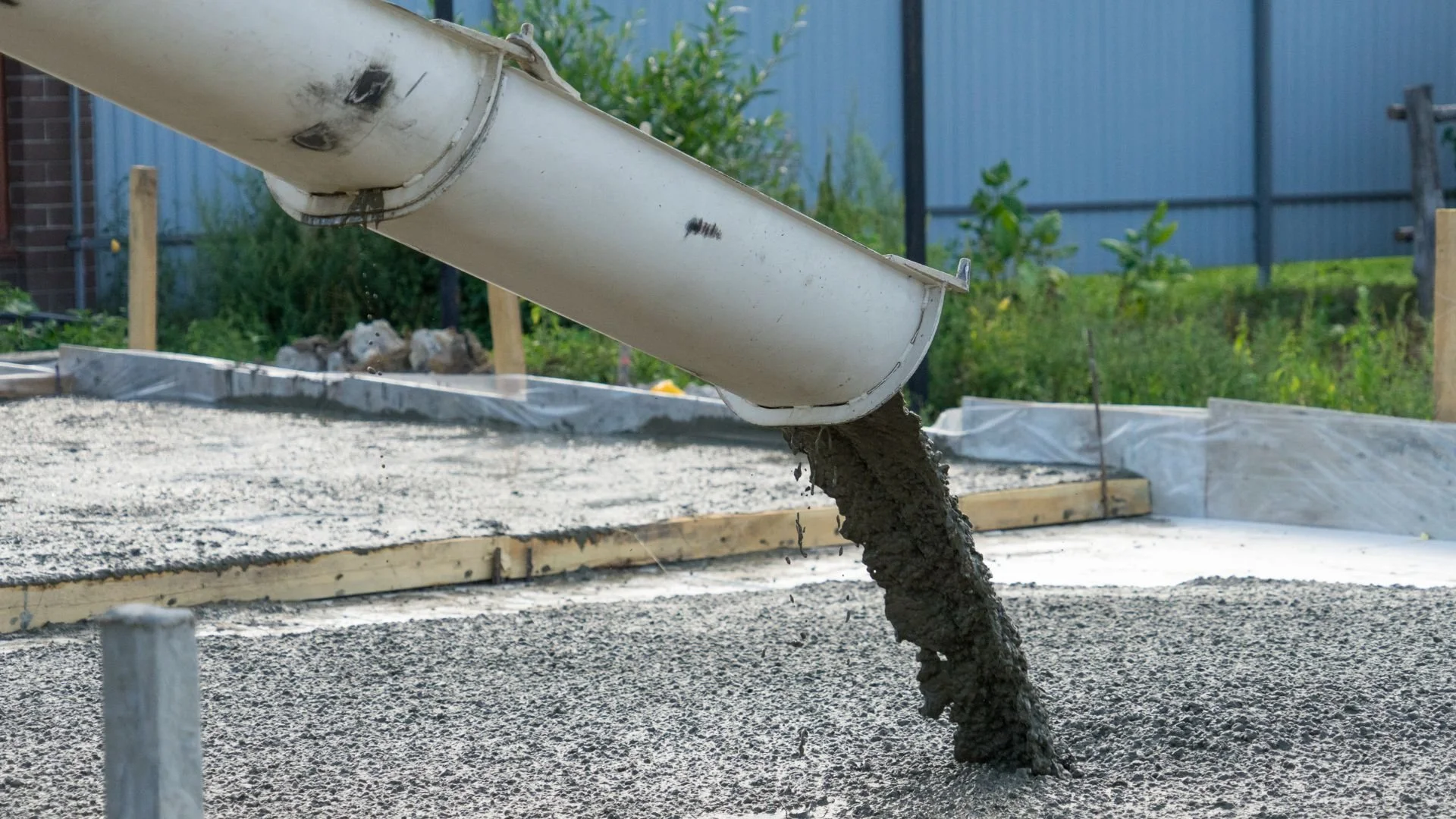 Concrete being poured from a white chute onto a construction site with a wooden frame, surrounded by fenced green area and garden plants in the background.