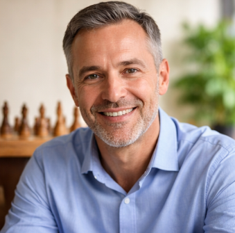 Smiling middle-aged man with gray hair and beard, wearing a blue shirt, indoors with blurred bookshelf and plant in the background.