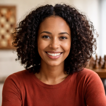 A woman with curly hair smiling, wearing a rust-colored sweater in a warmly lit room.