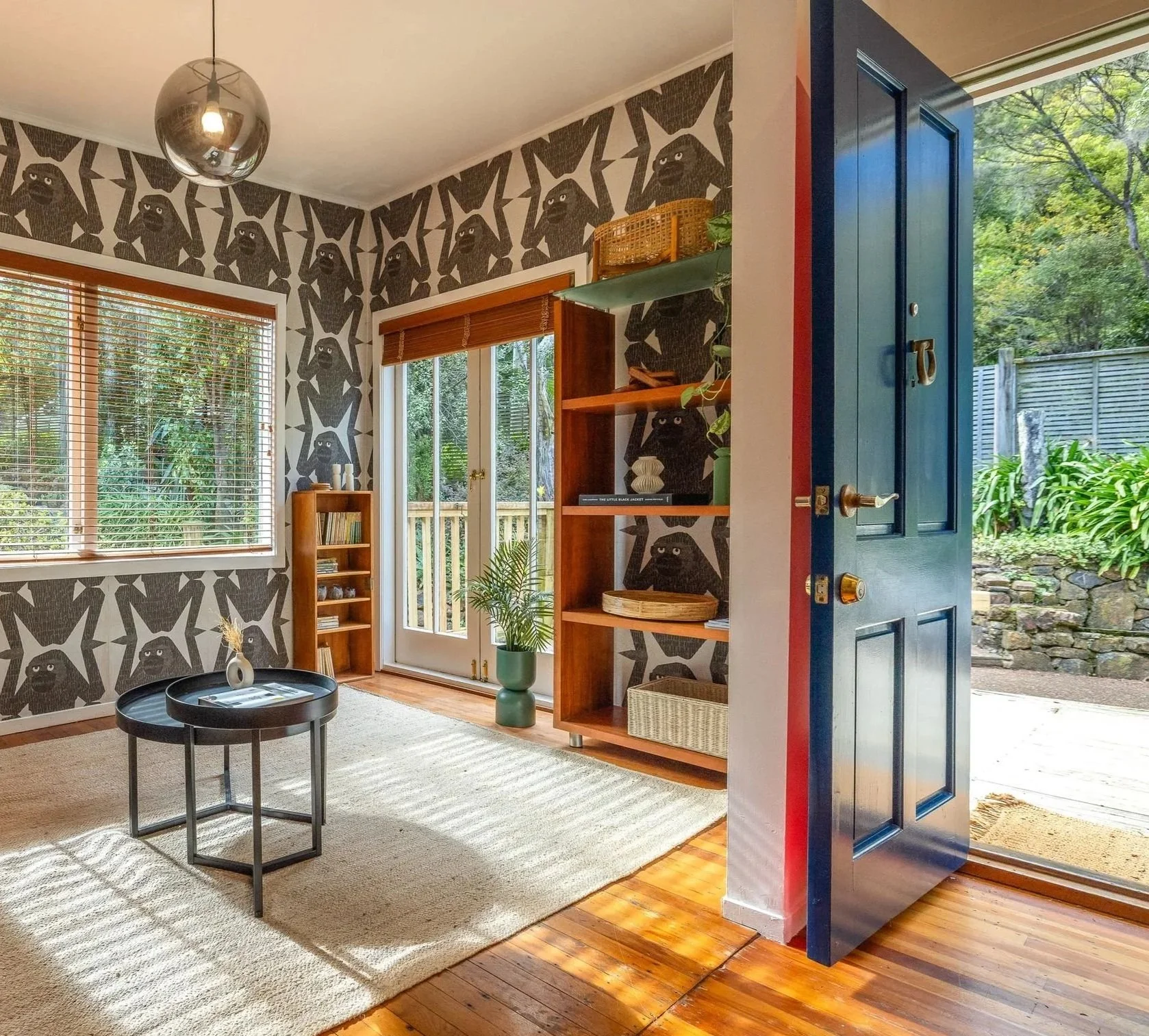 Interior of a room with patterned wallpaper featuring black dog's face designs, large window with blinds, glass sliding door with wooden frame leading outside, open blue door revealing exterior greenery, wooden shelf with decorative items, plush carpet, small round black table with decorative object, and potted houseplant.
