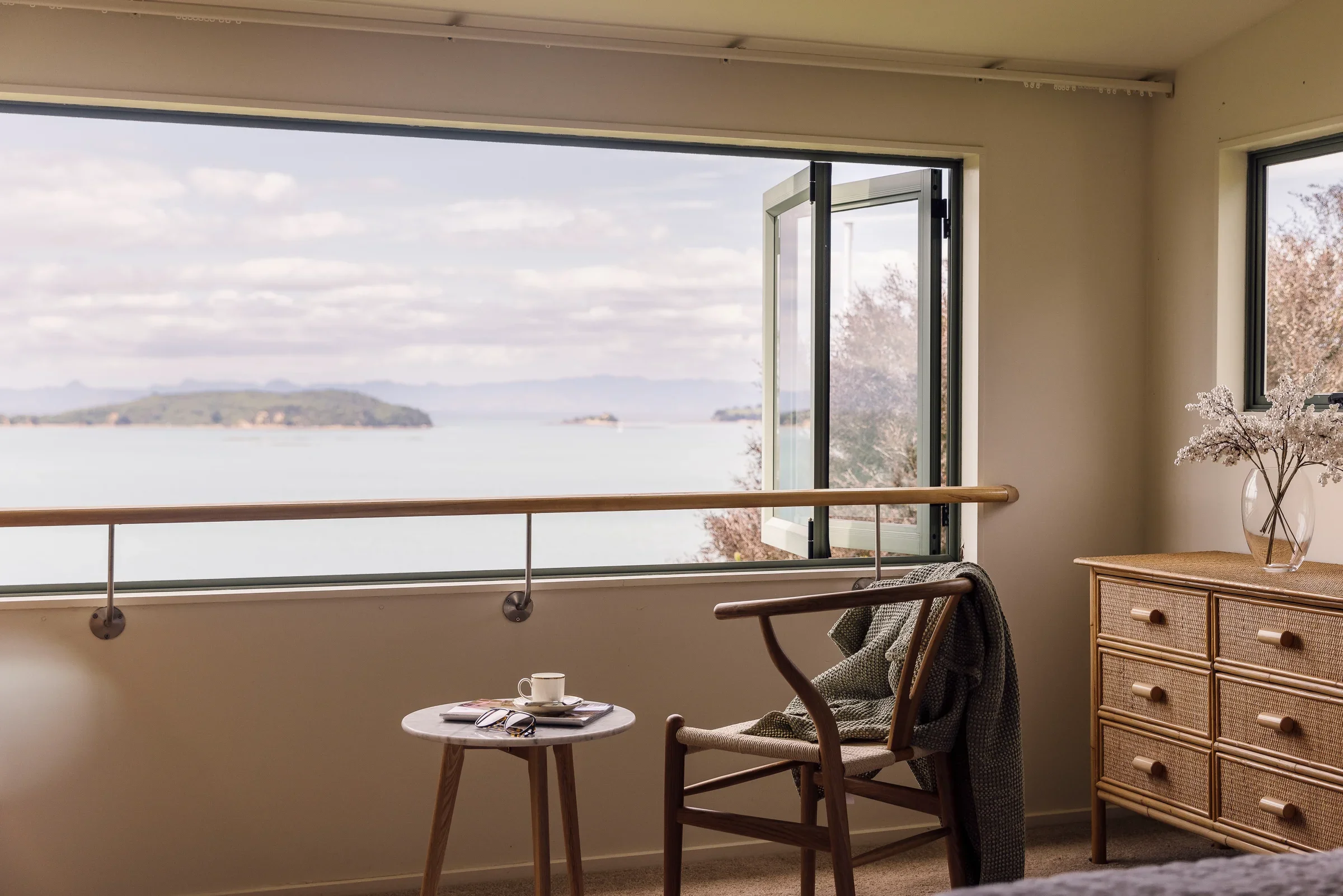 A room with an open window overlooking a body of water and distant islands, with a small round table holding a coffee cup, a spoon, and a book, along with a wicker dresser and a wooden chair.