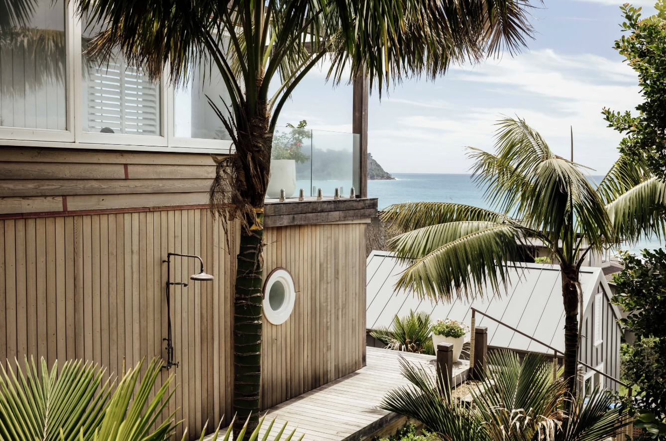 Outdoor shower area with wooden walls, surrounded by tropical palm trees, overlooking the ocean and a distant island in the background.