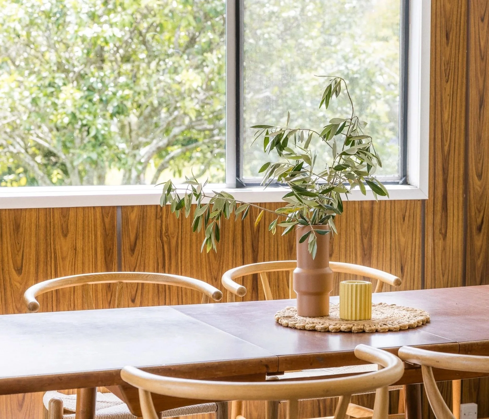 A dining room table with a flower vase centerpiece, yellow candle, placemat, and chairs, with a window showing green trees outside.