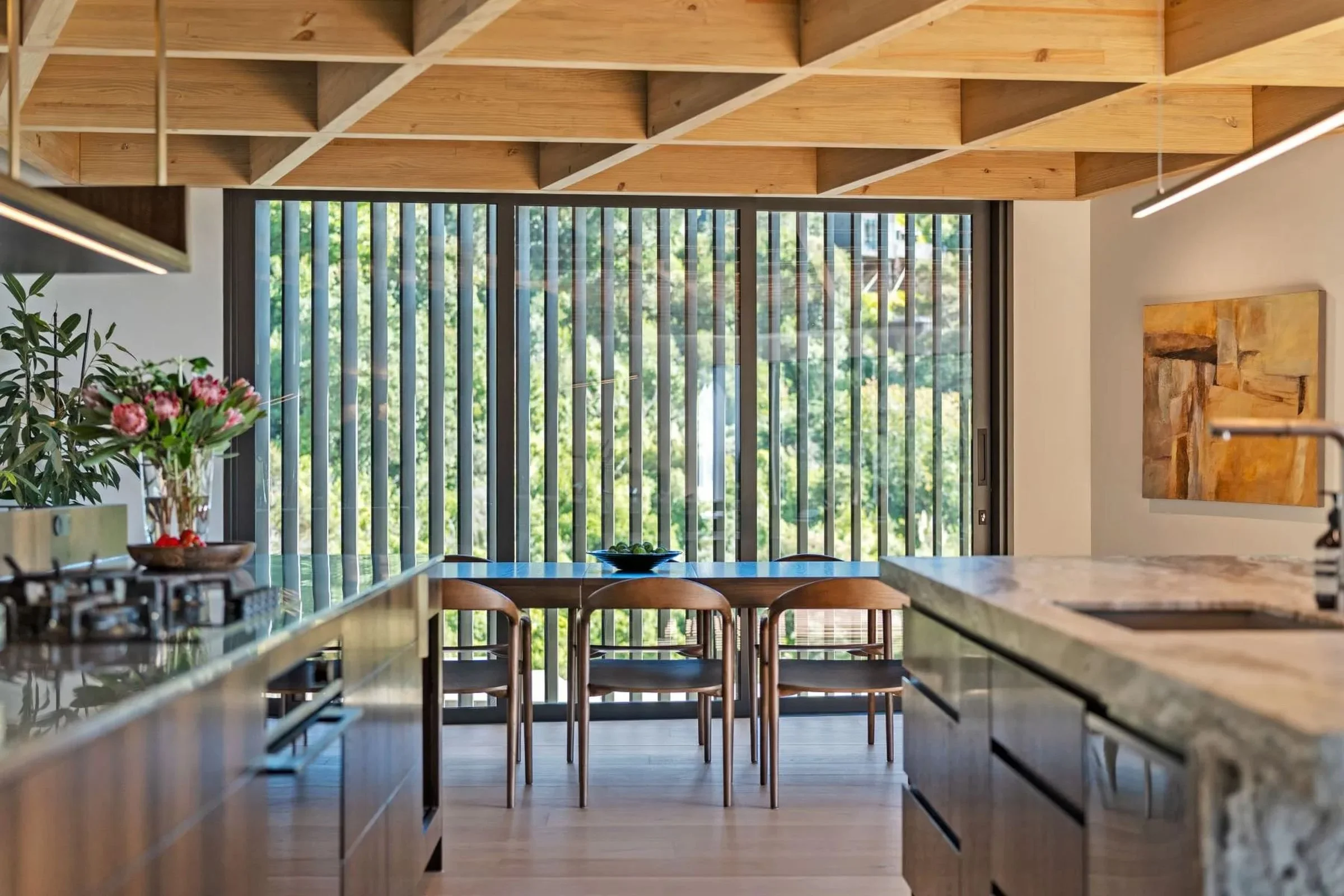 Modern kitchen interior with a large window and vertical blinds, wooden ceiling panels, a granite countertop, and a vase with pink flowers.