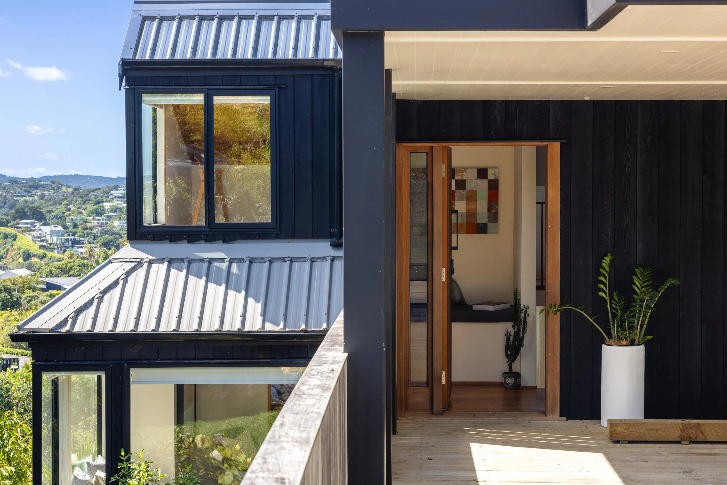 Modern black house with large windows, a metal roof, and a balcony with potted plants and art displayed inside, overlooking a scenic hillside.