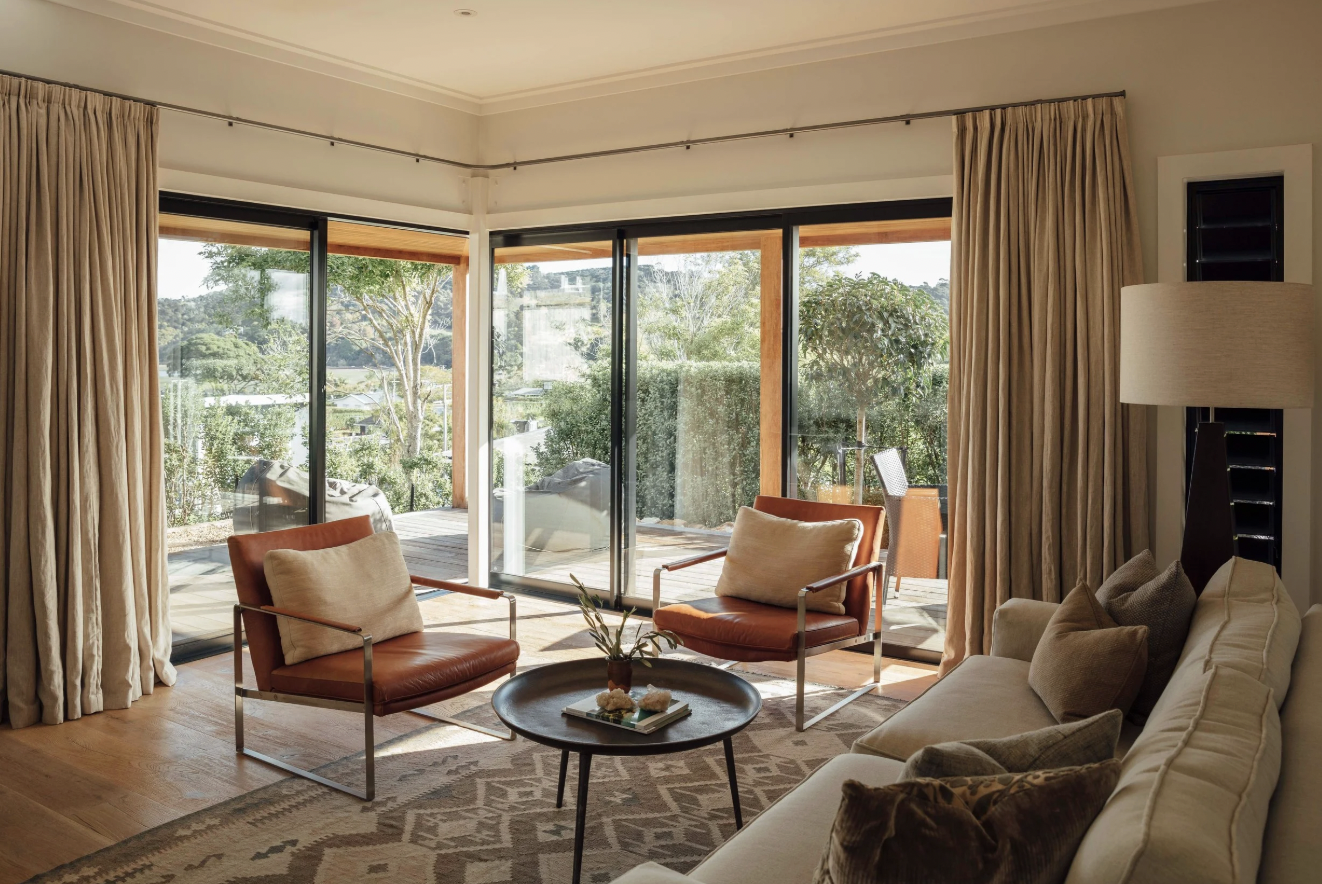 Living room with large sliding glass doors, beige curtains, a white sofa with beige and brown cushions, two brown leather armchairs with beige pillows, a round coffee table with a plant, and a view of a patio with furniture and greenery outside.