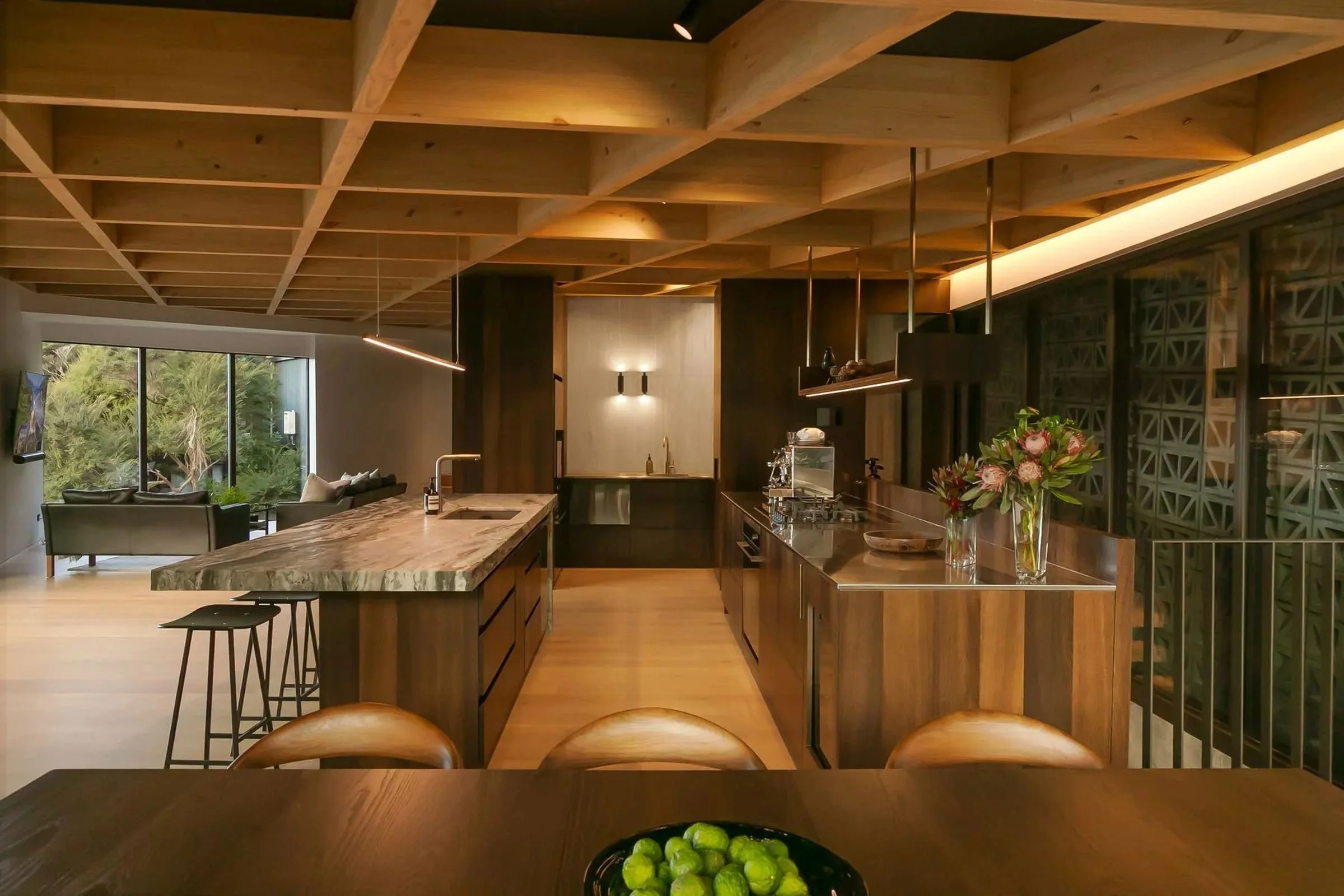 Modern kitchen with wooden ceiling beams, large kitchen island, and a dining table with seats in the foreground. There are decorative elements such as a vase with flowers and a bowl of green apples on the table.