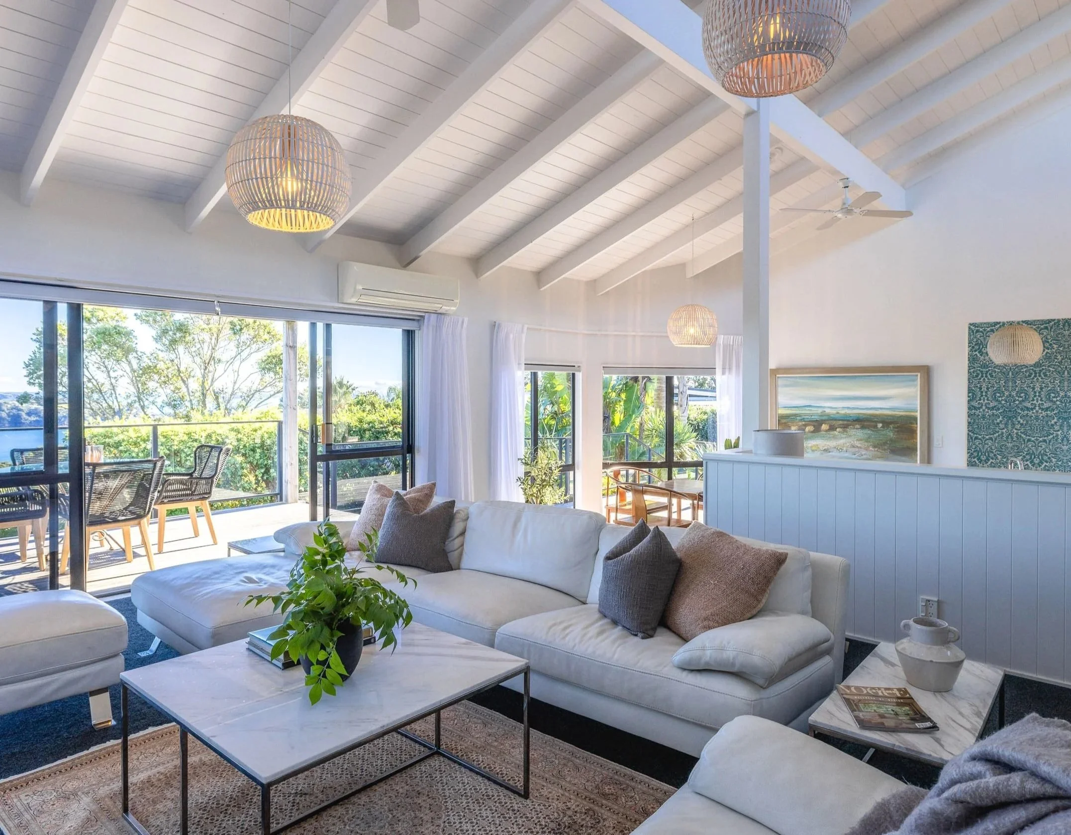 Bright interior living room with white sofa, green plants, marble coffee table, large windows, and ceiling with wooden beams and hanging woven light fixtures.