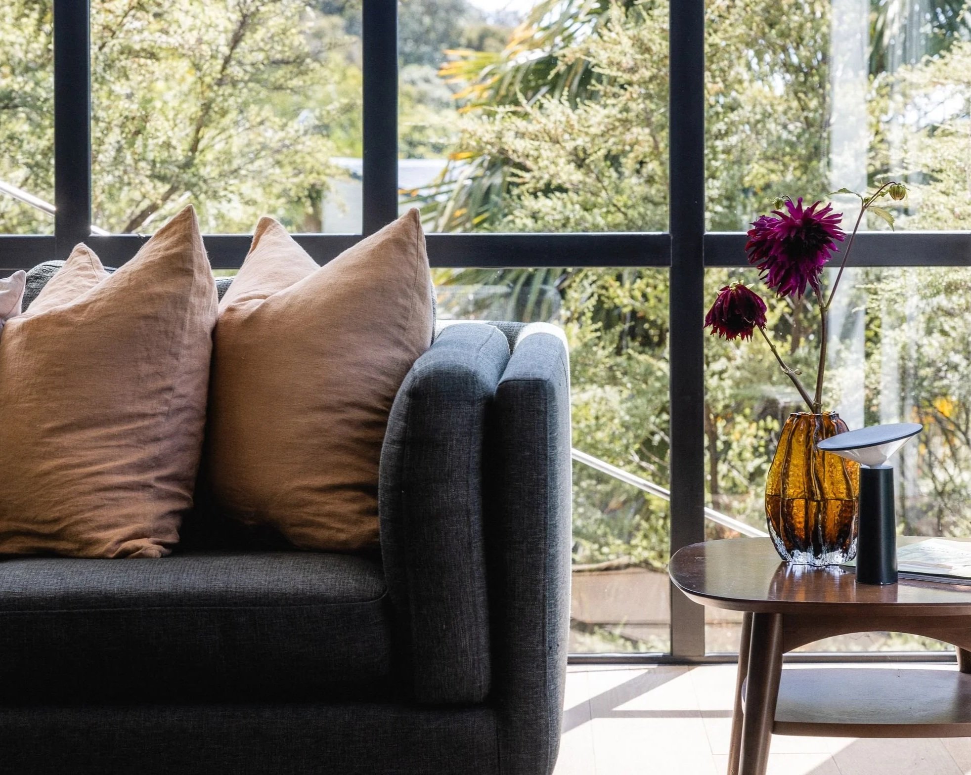 Living room with cushions on a sofa near large glass window revealing greenery outside, and a side table with a decorative glass vase and a book.
