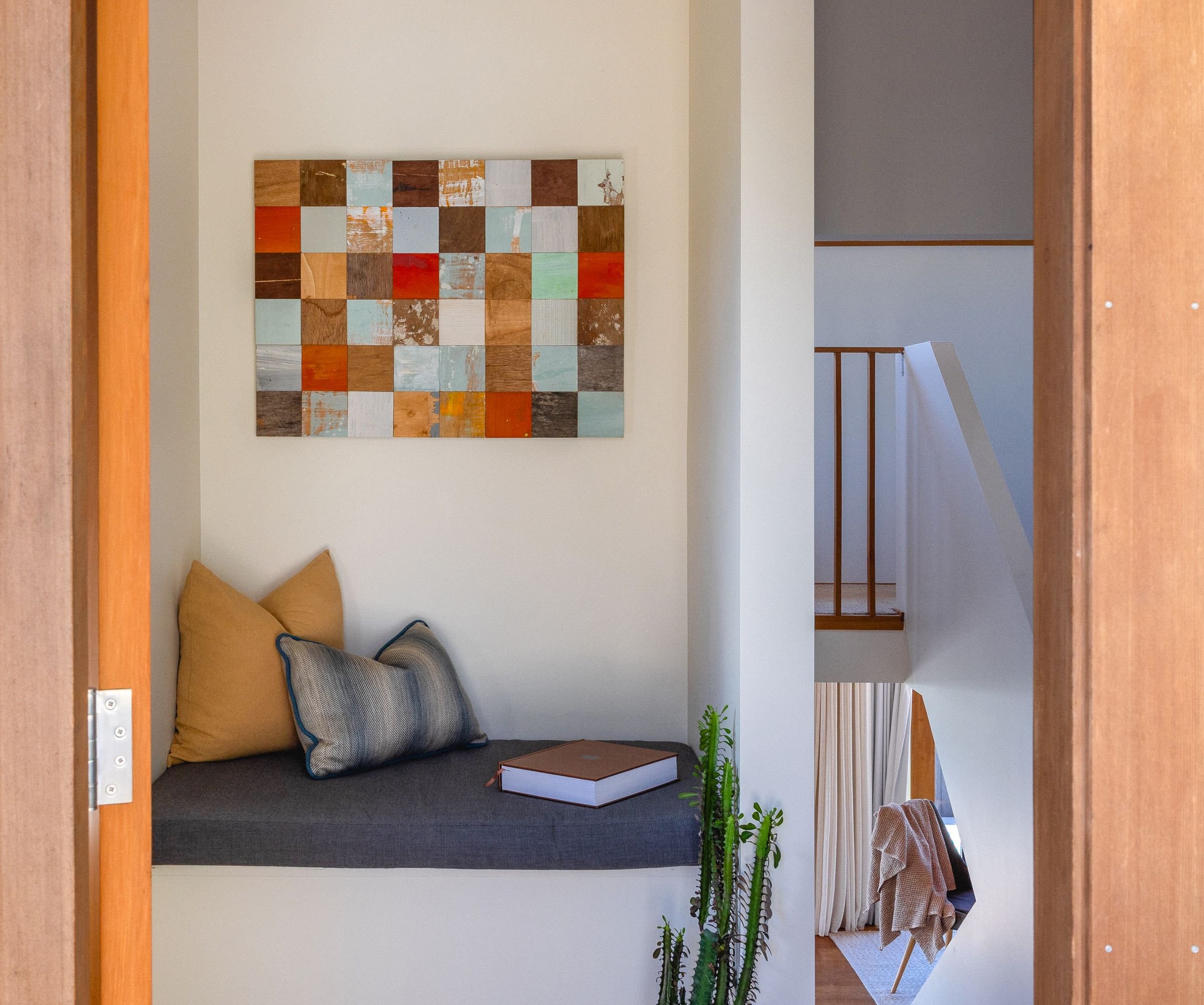 View of a cozy seating nook with a dark couch, a beige throw pillow, a large book, green cactus plants, and a wall art piece with a grid of colorful, textured wooden squares. The nook is on a platform with a railing and white curtains on the right side.
