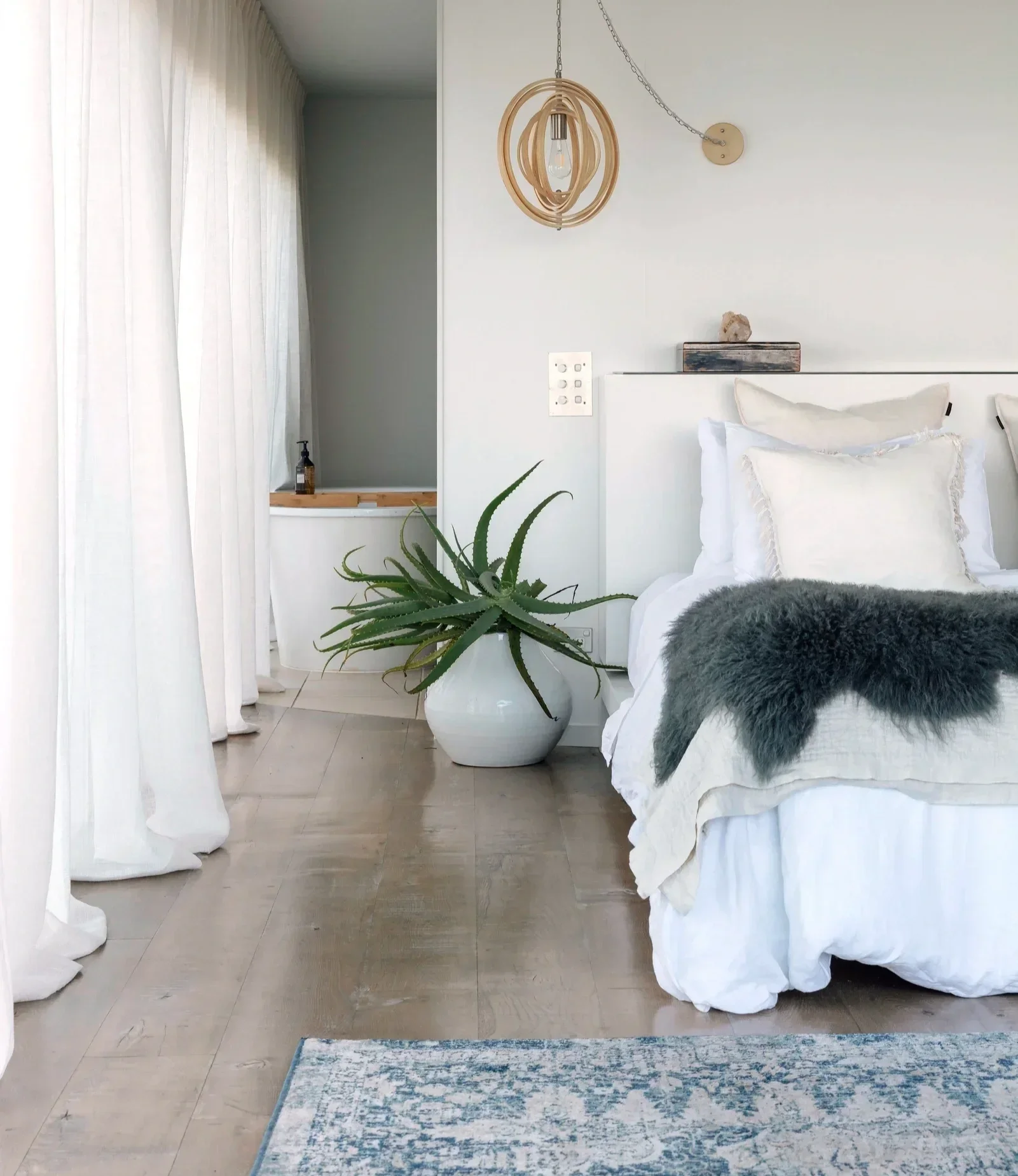 A bedroom with white walls and light wood flooring, decorated in a minimalist style. There is a bed with white bedding and a dark gray furry blanket. Next to the bed is a large white vase with a green aloe plant. To the left, sheer white curtains cover a window, and a small wooden ledge with a bottle of soap or lotion is beneath it. A modern, wooden pendant light hangs from the ceiling. On the wall behind the bed is a small piece of decor, possibly a rock or mineral, in a wooden box. There is a patterned rug in front of the bed.