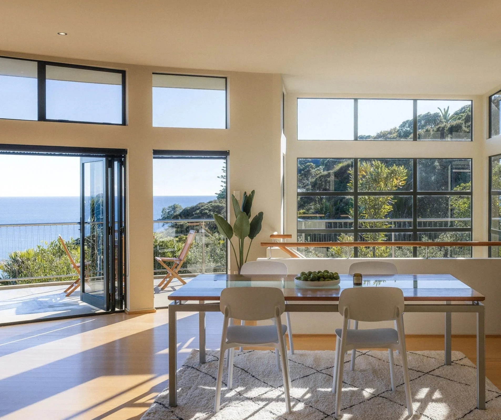 Sunlit dining area with large floor-to-ceiling windows offering ocean and greenery views, wooden dining table with white chairs, potted plant, and sliding glass door leading to a balcony with outdoor chairs.