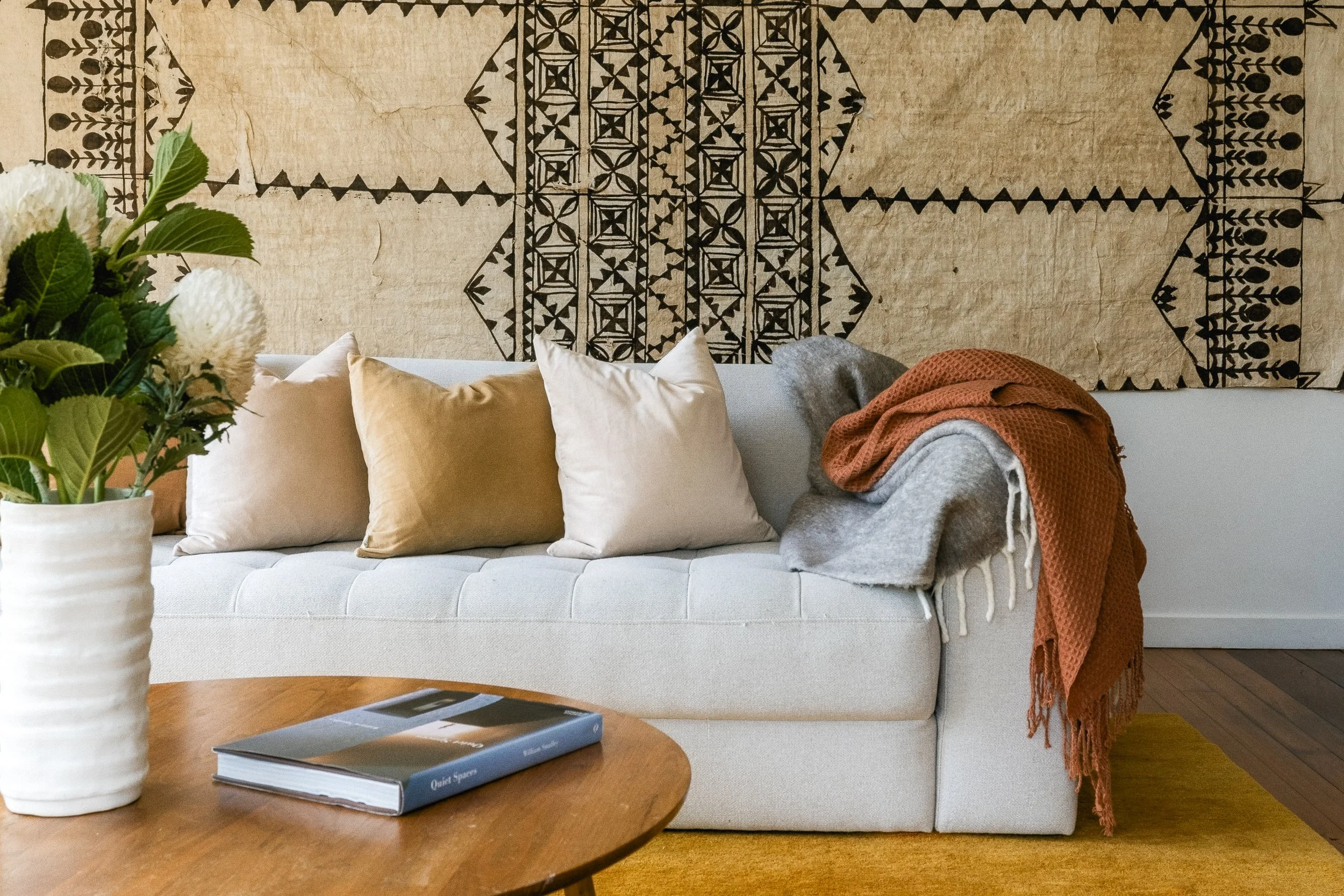 Living room with a white sofa, beige and white pillows, a gray blanket, a wooden coffee table with a book, and a large wall hanging with black geometric and plant patterns. A plant with white flowers is partially visible on the left.