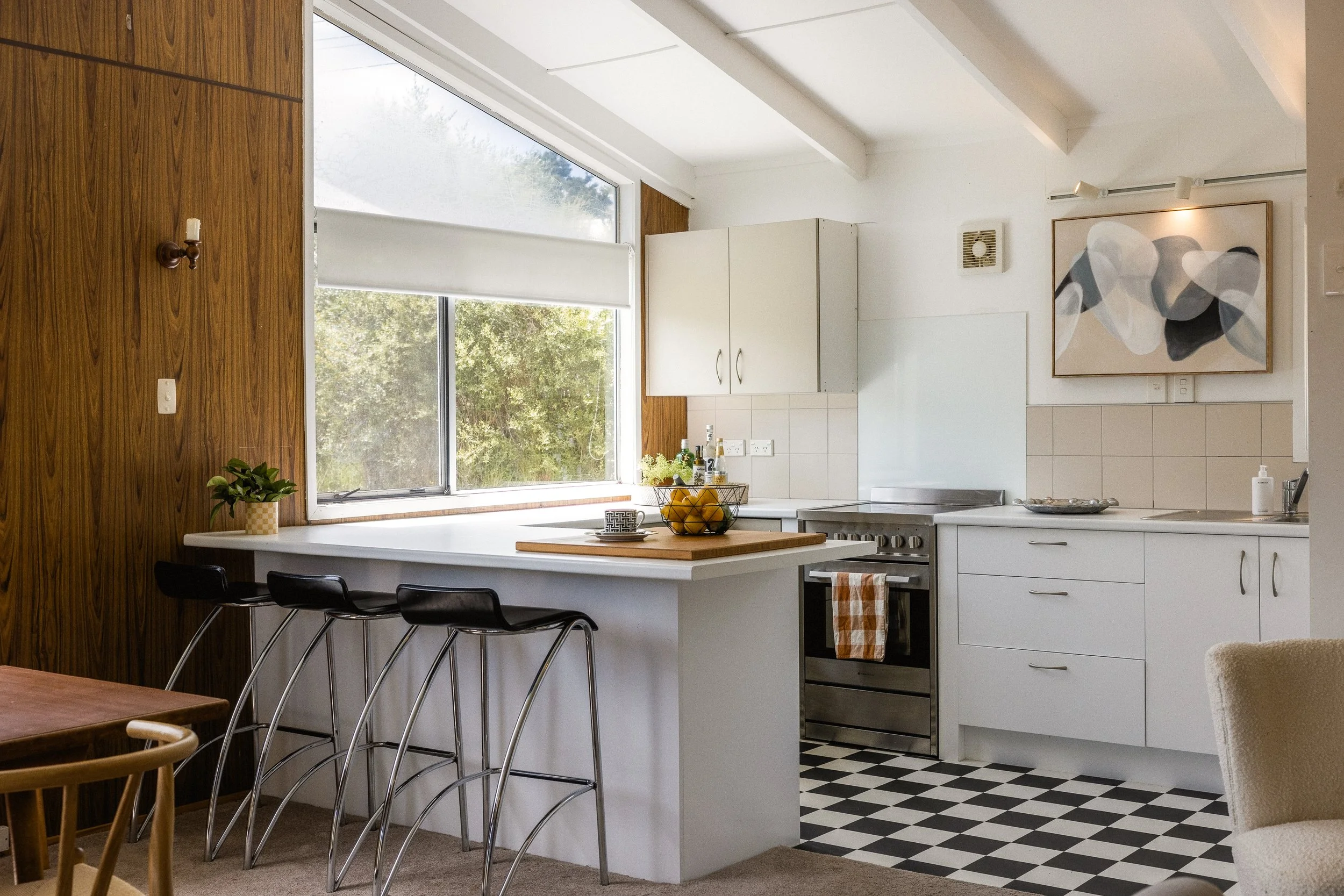 A bright kitchen with white cabinets, a black and white checkered floor, and a large window. A kitchen island with a wooden cutting board, a bowl of lemons, and some bottles. Modern art hangs on the wall.