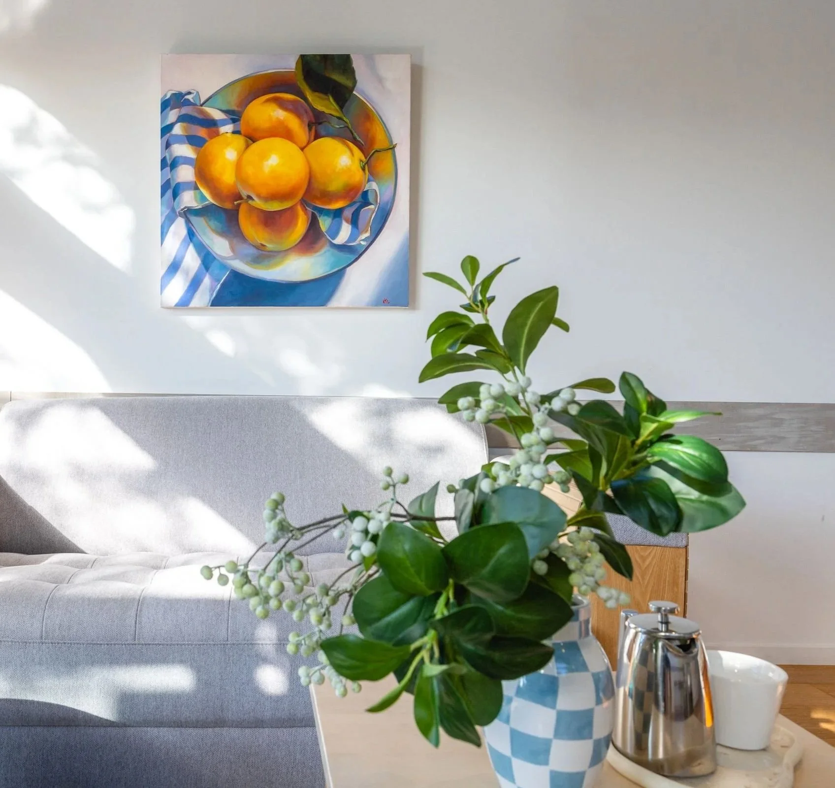 Decorative kitchen countertop with a green leafy plant with white berries in a blue and white checkered vase. Behind, a silver soap dispenser and white cup, with a light gray cushioned bench and a wall-mounted still life painting of a bowl of oranges and a peach. Sunlight casting shadows across the scene.