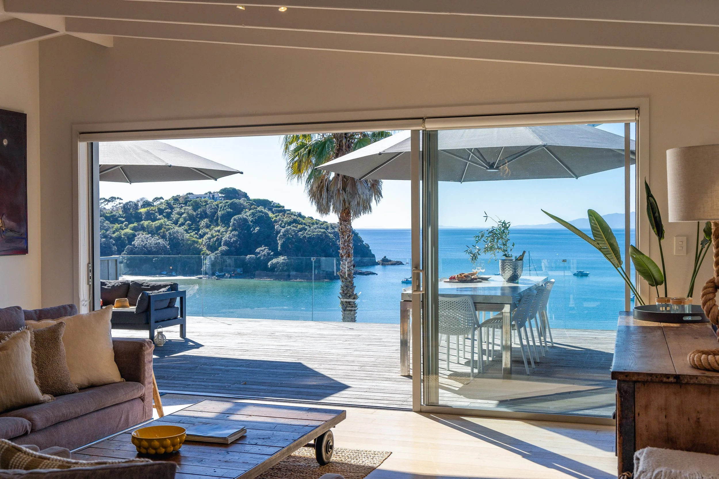 Interior of a living room with sliding glass doors opening to a balcony overlooking a beach, with umbrellas, palm trees, and ocean scenery in the background.