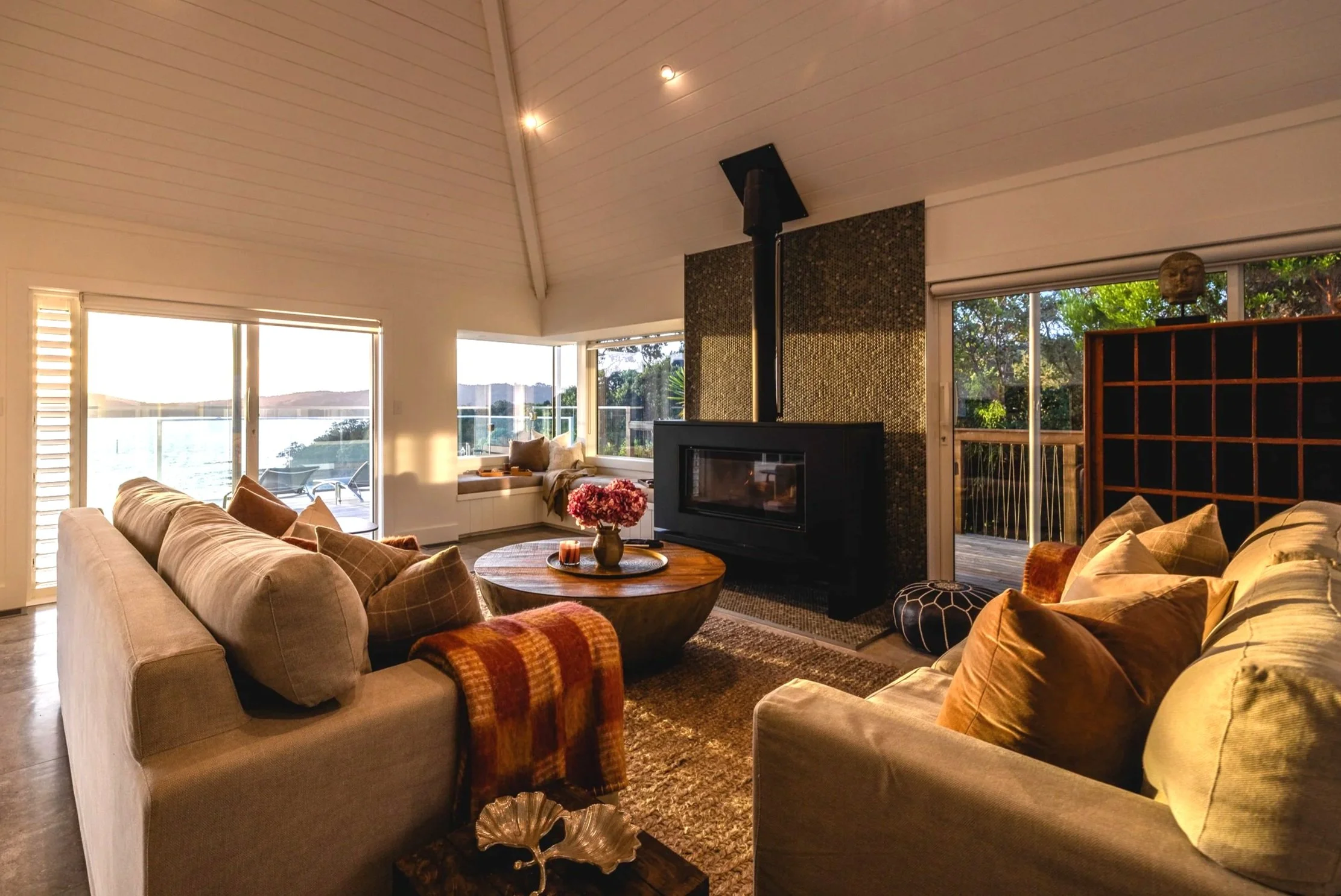 Living room with beige sofas, throw pillows, a round wooden coffee table with a vase of pink flowers, large windows showing a view of water and trees, fireplace, and ceiling lights.
