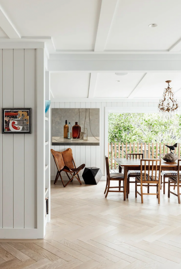 Bright dining room with wooden table and six chairs, a leather chair, decorative vases on a ledge, a chandelier, and a large window showing green foliage outside.