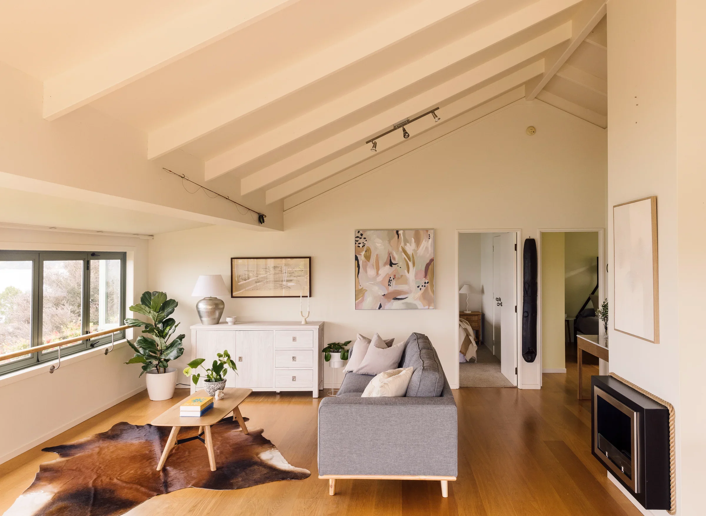 Living room with large window, potted plants, a couch, coffee table, artwork on the wall, and a ceiling with exposed beams.