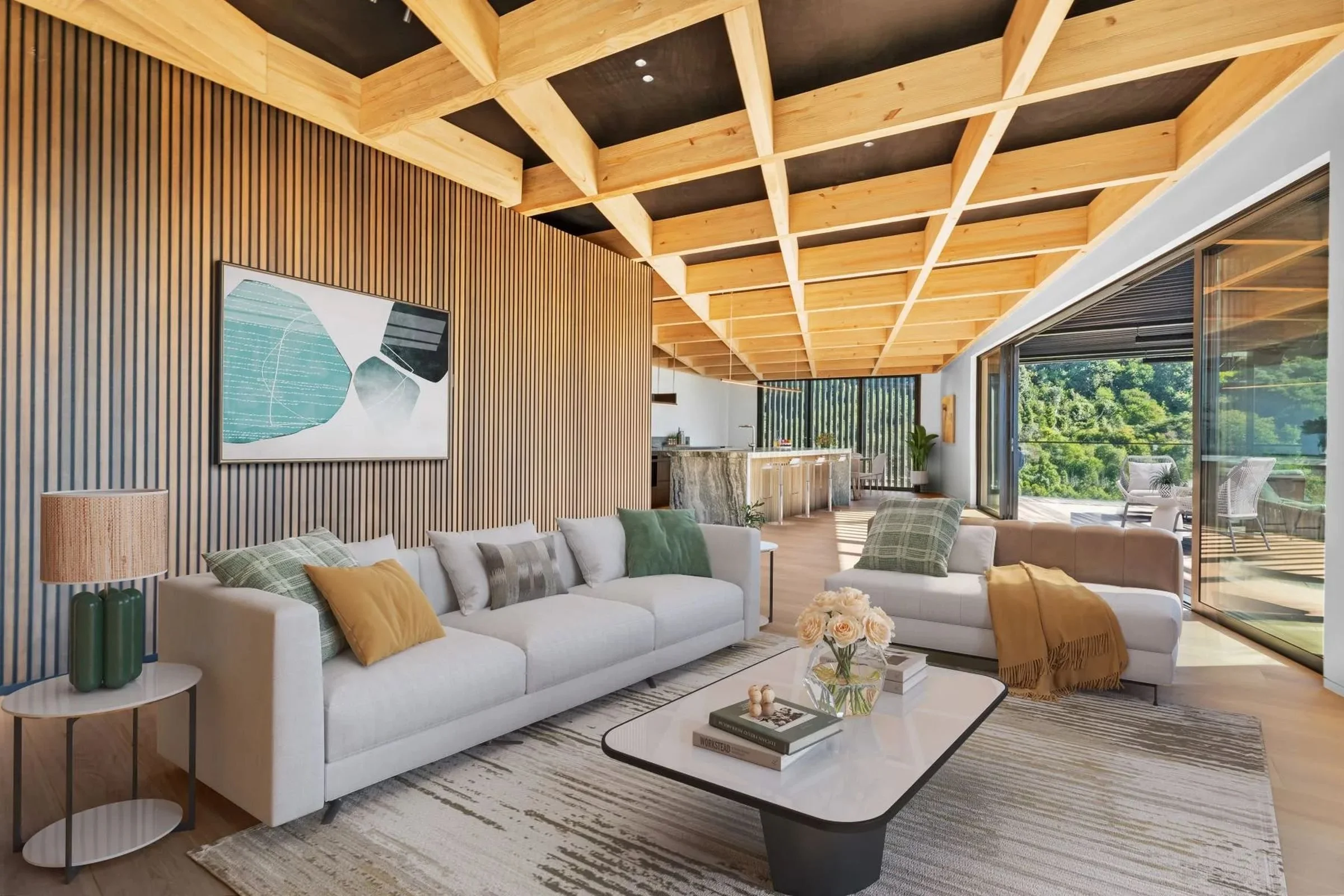 Modern living room with a white sofa, decorative pillows, a coffee table with books, a patterned rug, a side table with a lamp, wood paneled walls, an abstract painting, and a large window showing a balcony with outdoor furniture and greenery.