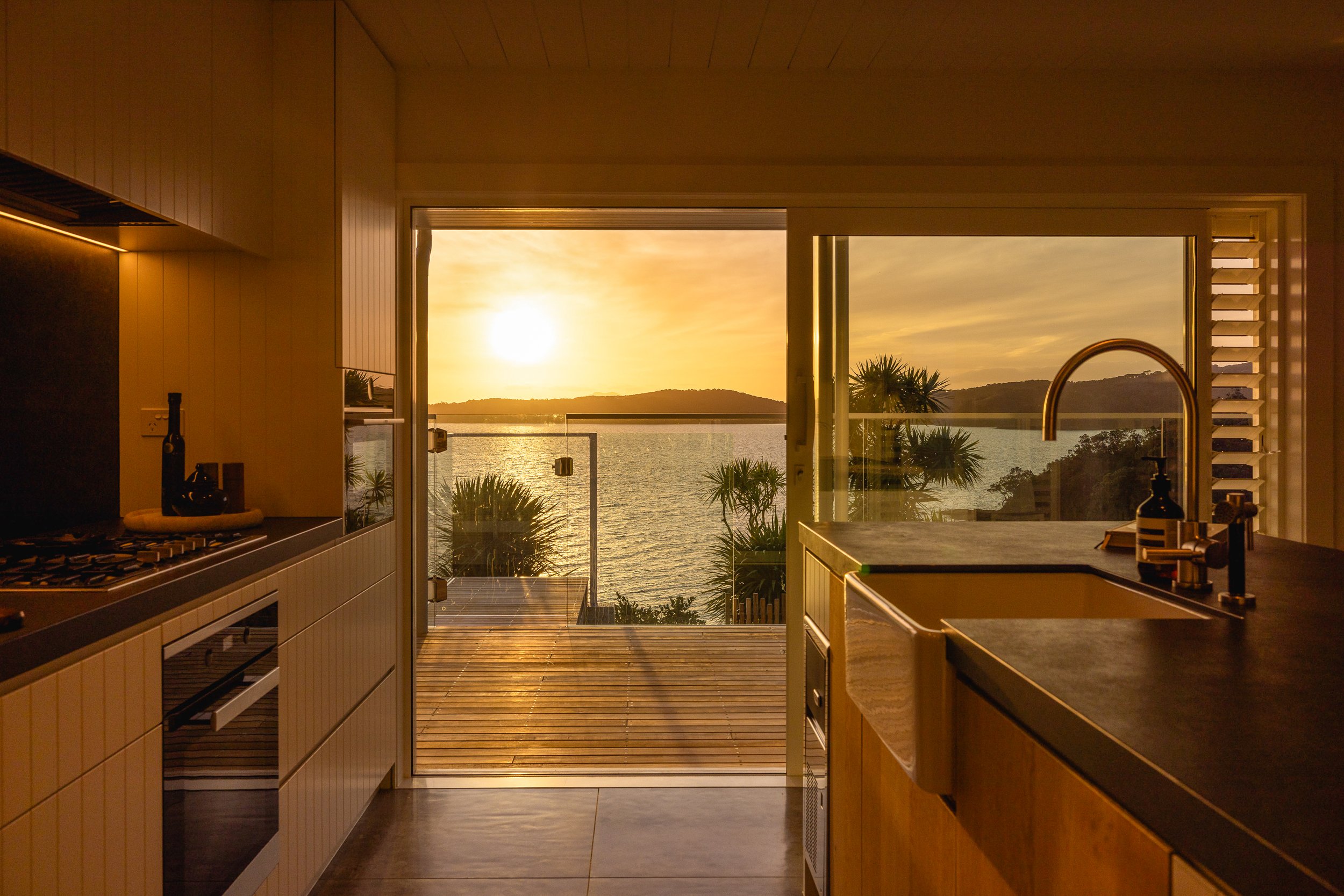 Sunset view through open kitchen doors, overlooking a water body with hills and palm trees in the distance.