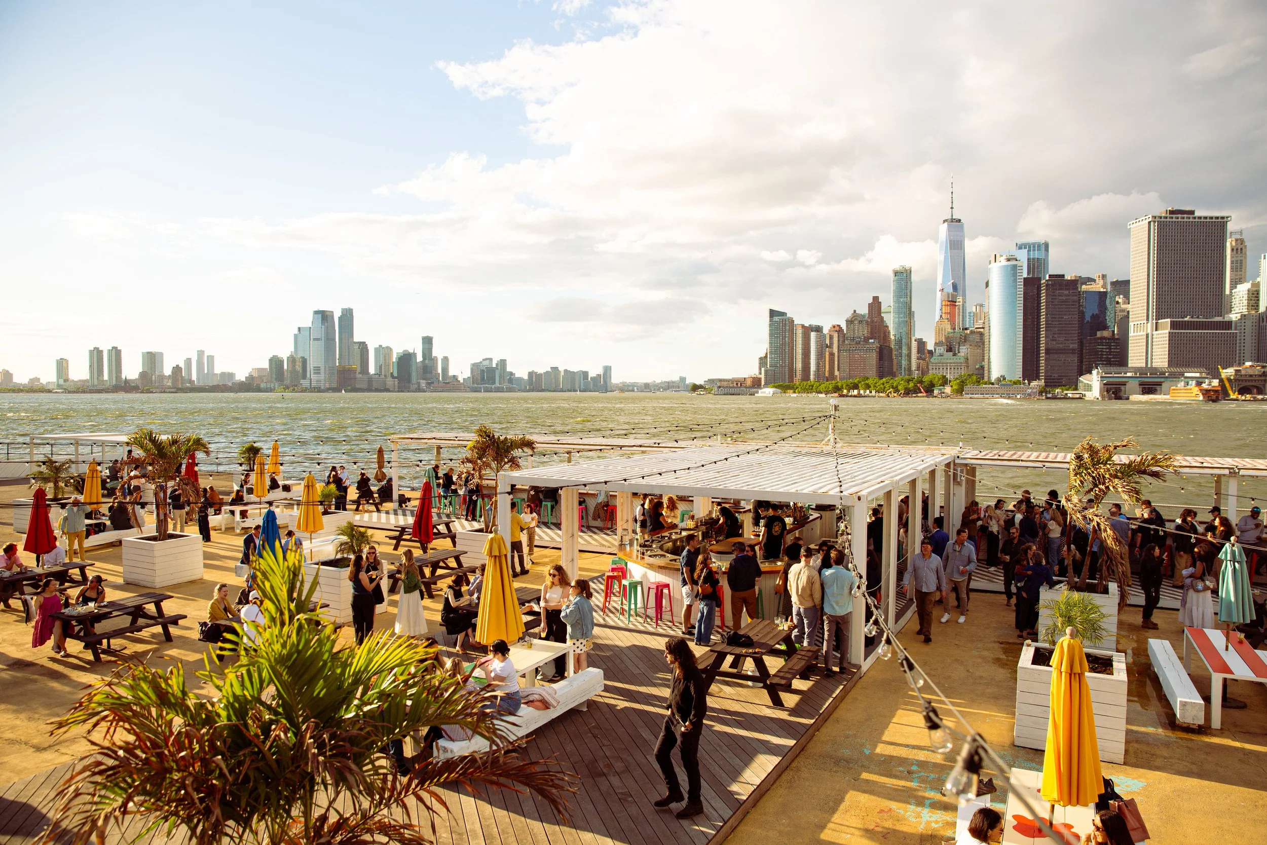 Crowded outdoor waterfront bar, Six Coasts on Governors Island, with patrons at tables under a white pergola and a panoramic view of the Lower Manhattan skyline across the water.