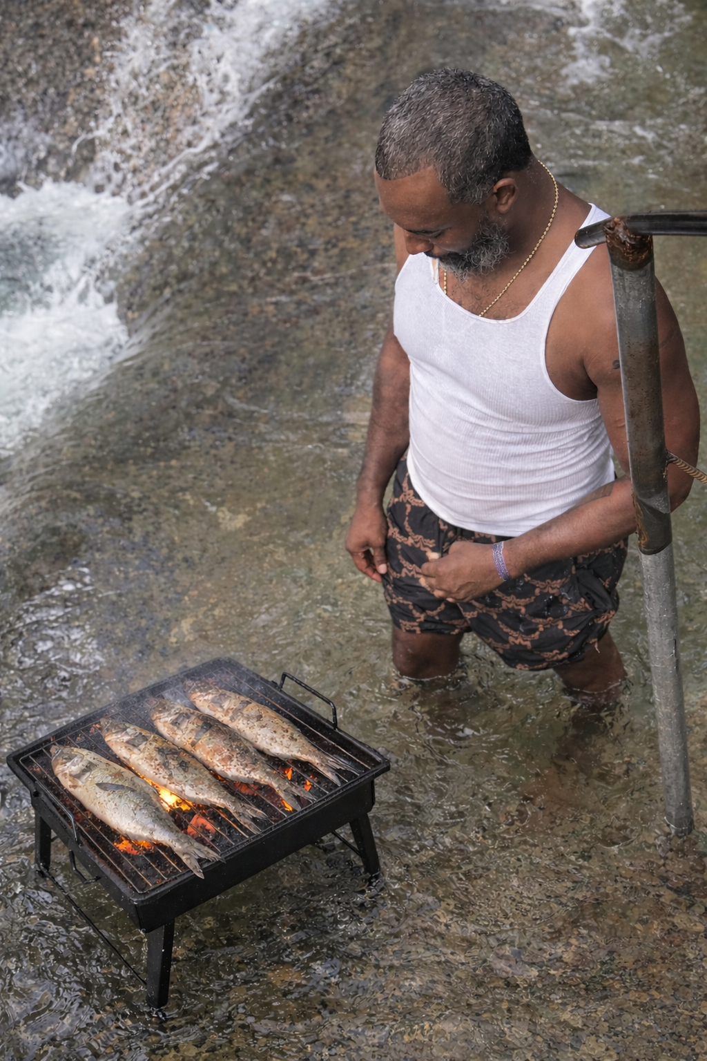 A man standing in shallow water near a rocky riverbank, grilling fish on a portable grill placed on the water, while looking down at the fish.